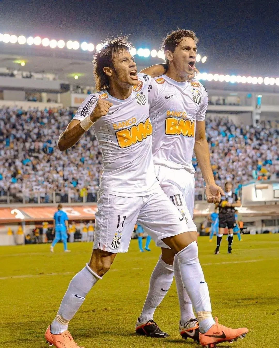 Two soccer players celebrating on the field during a night match, wearing white uniforms with logos and sponsorships, and cheering with arms around each other.