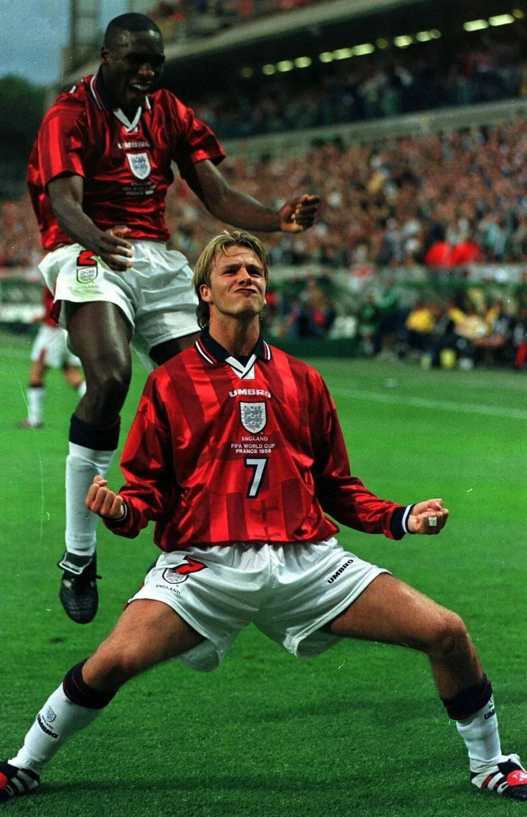 Two soccer players celebrating on the field during a match. One player is jumping, and the other is kneeling with fists clenched, both wearing red England jerseys from the 1998 FIFA World Cup.