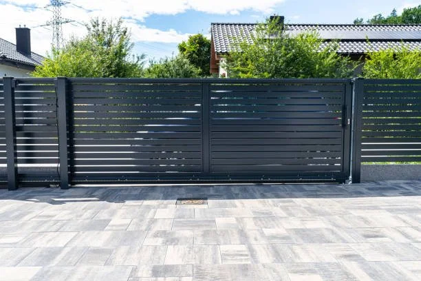Black horizontal slat fence with a gate, on a paved driveway, with trees and houses in the background under a blue sky.
