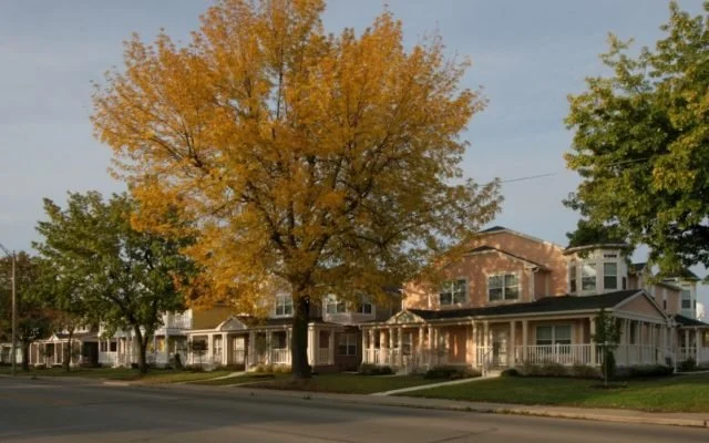A row of houses on a residential street with trees, one tree with yellow leaves, and a partly cloudy sky.