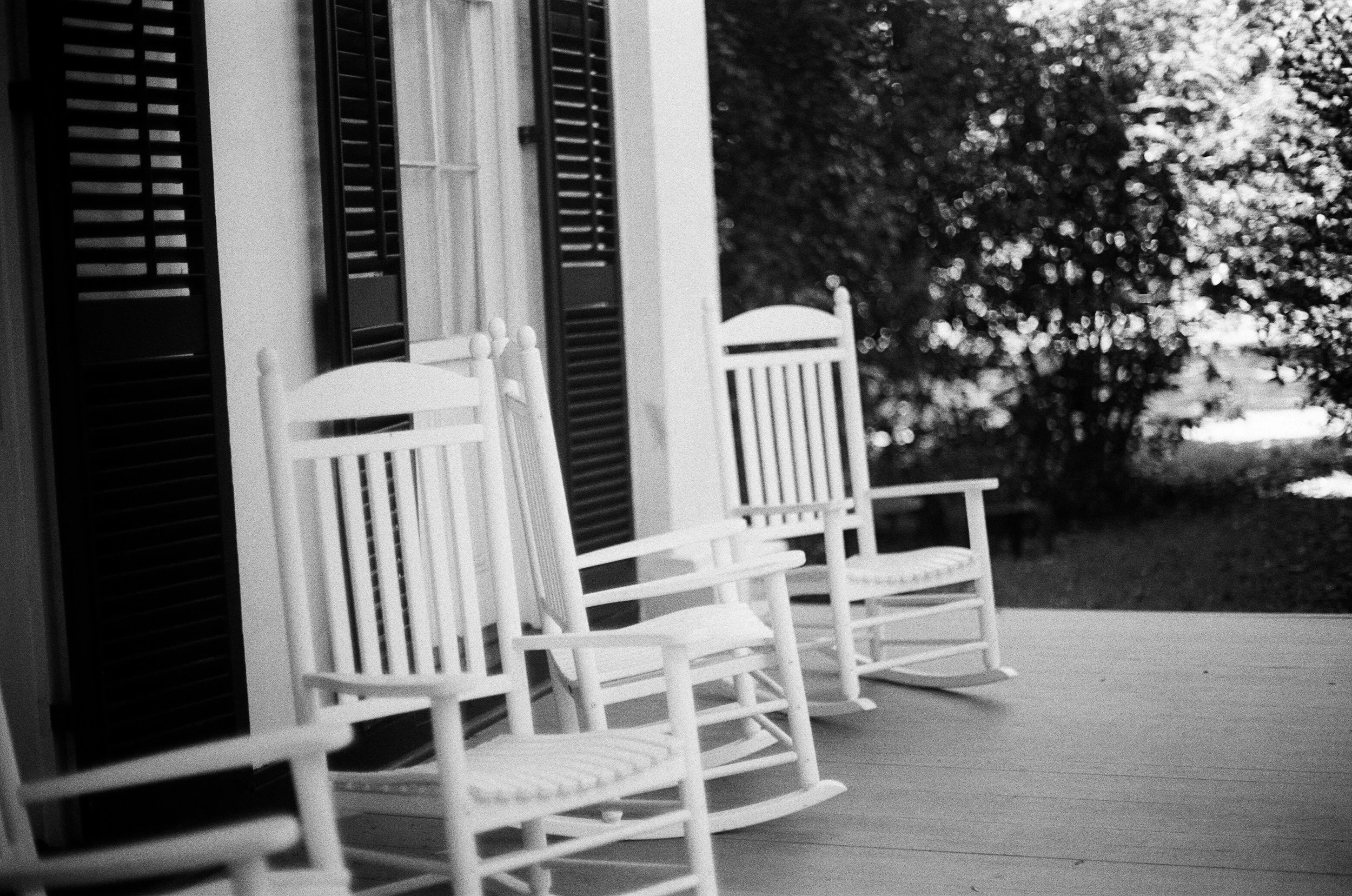 Black and white photo of three rocking chairs on a porch, with a house featuring shutters and trees in the background.