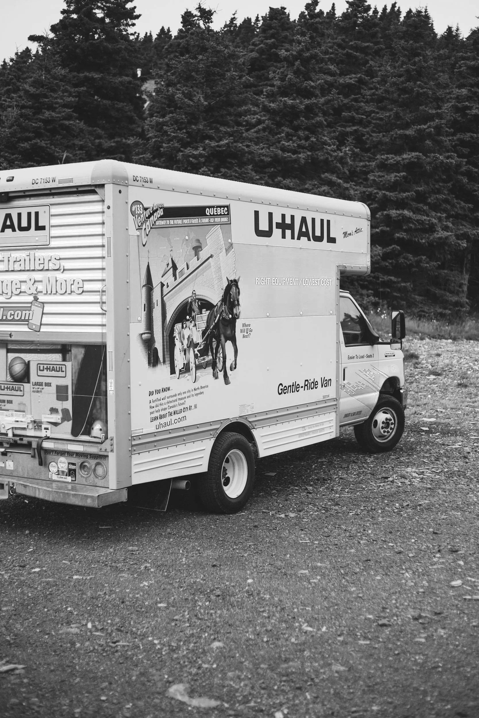 A black and white photo of a U-Haul moving truck on a gravel surface with a background of trees.