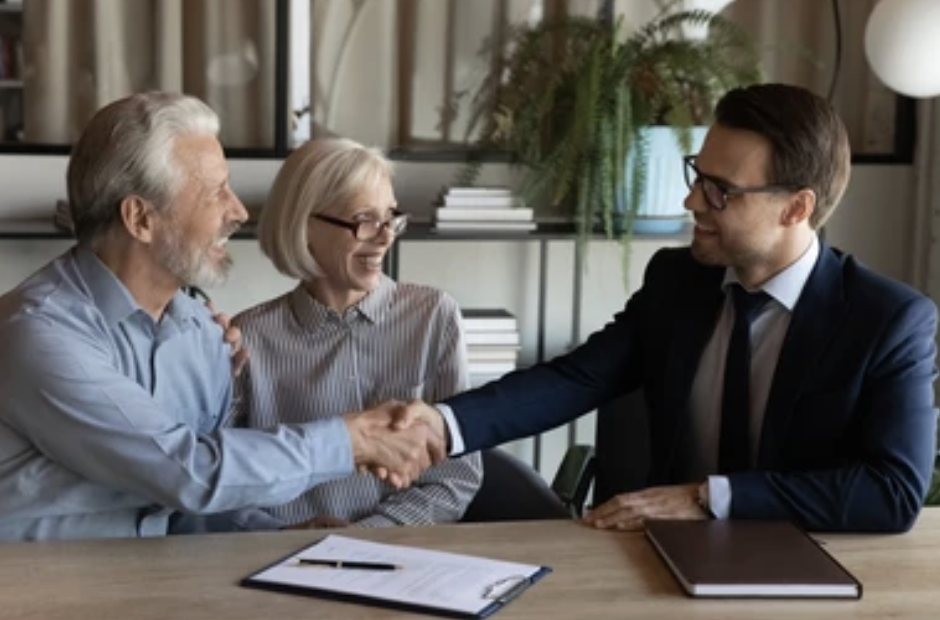 A young man in a business suit shaking hands with an older man in a light blue shirt, while an older woman with glasses looks on and smiles in an office setting.