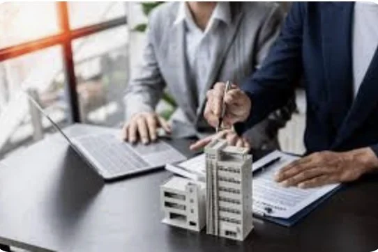 Two people in business attire discussing building models and documents at a desk.