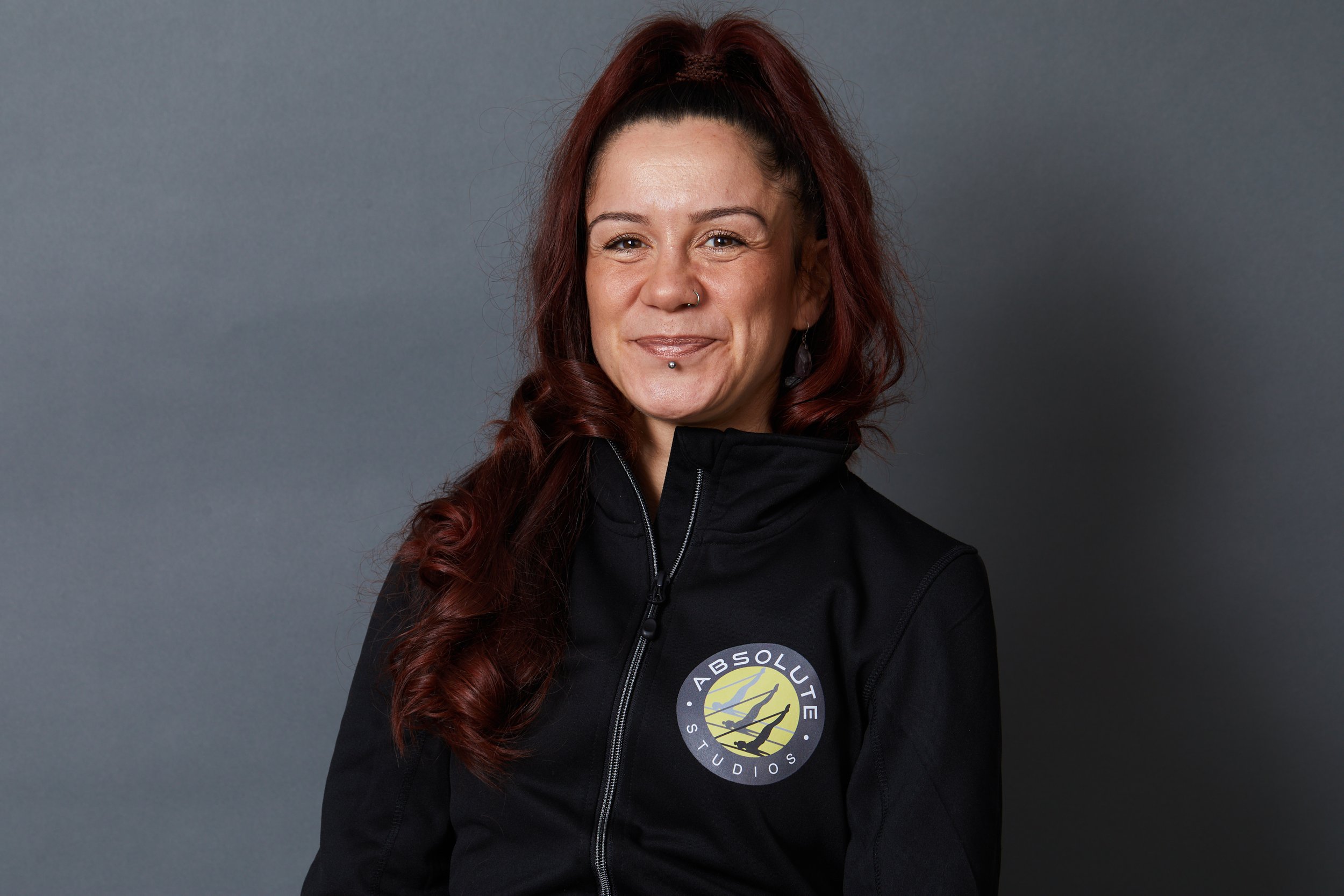 A smiling woman with long, wavy, red hair in a high ponytail, wearing a black jacket with a logo for Absolute Studios, standing against a gray background.