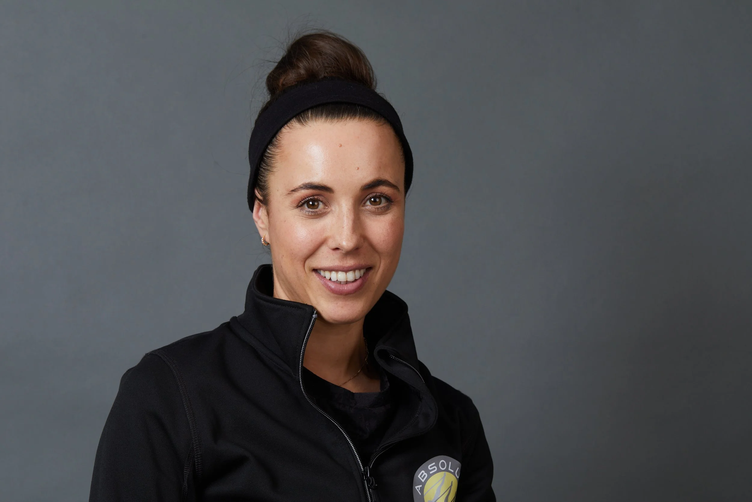 A woman with a high bun hair tied with a black headband, wearing a black zip-up jacket with a logo, smiling at the camera, standing against a gray background.