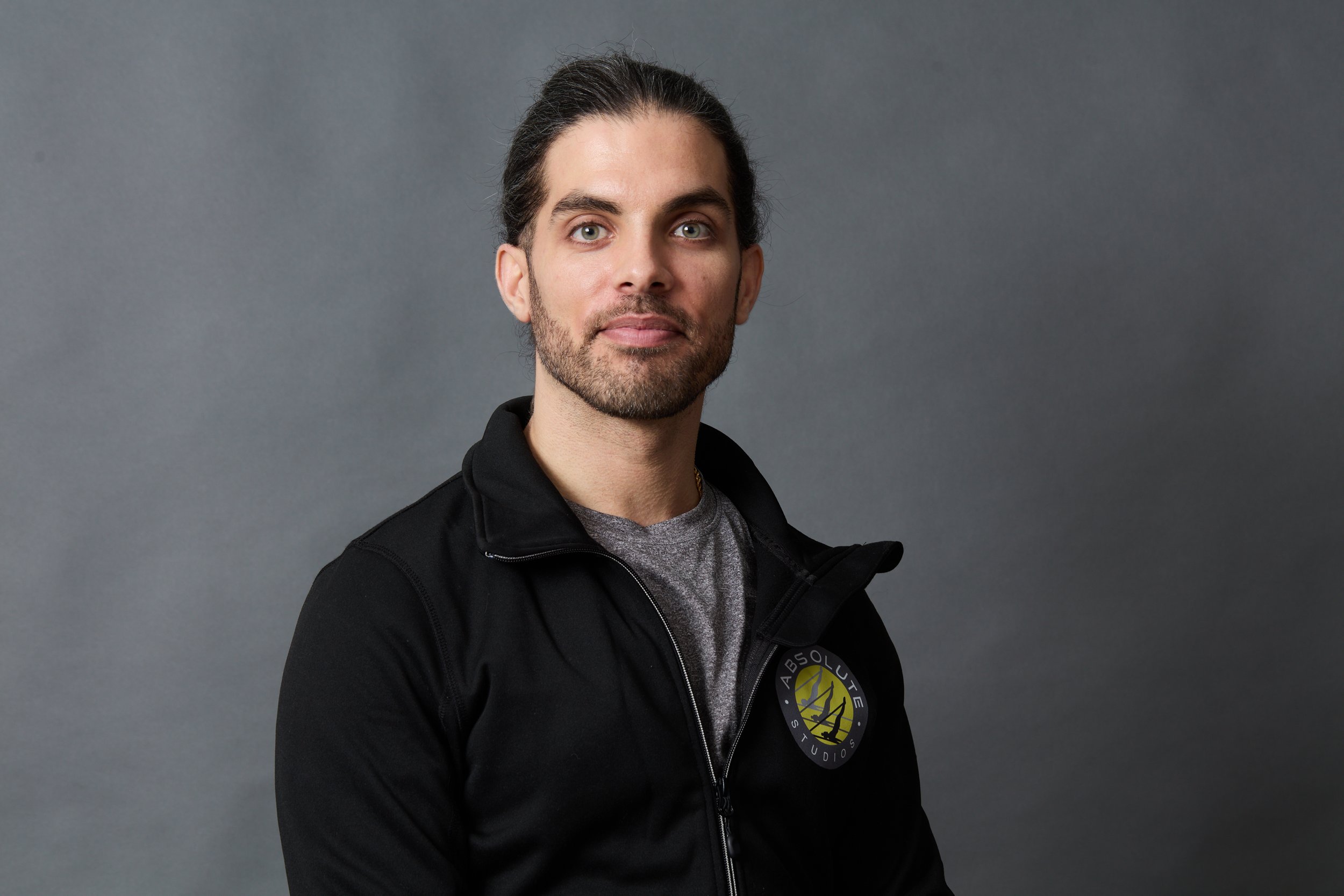 Portrait of a man with long dark hair, beard, wearing a black zip-up jacket with a logo, and a gray shirt, against a gray background.