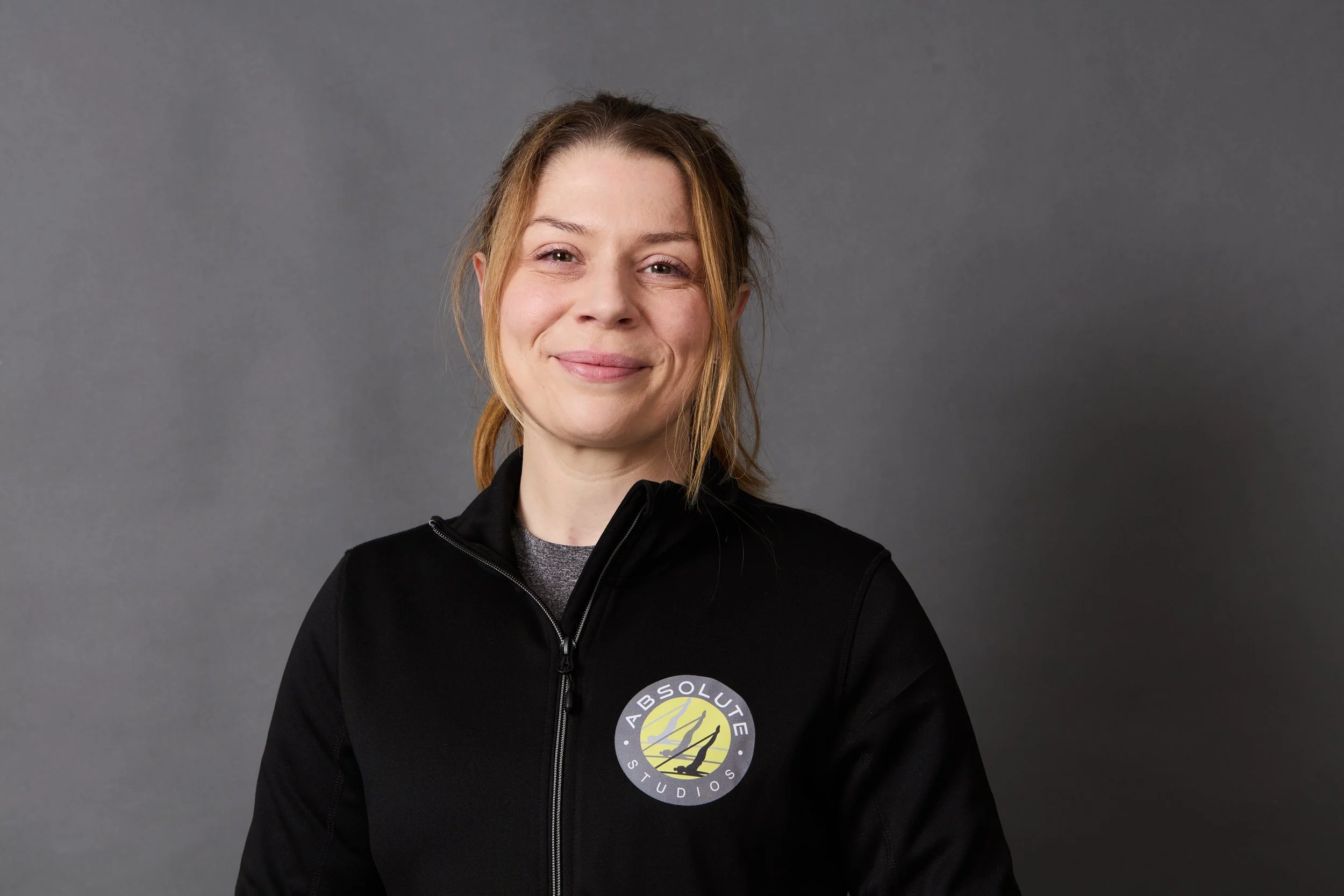 A young woman with blonde hair wearing a black jacket with a logo reading 'Absolute Studios' on the chest, standing against a gray background, smiling.