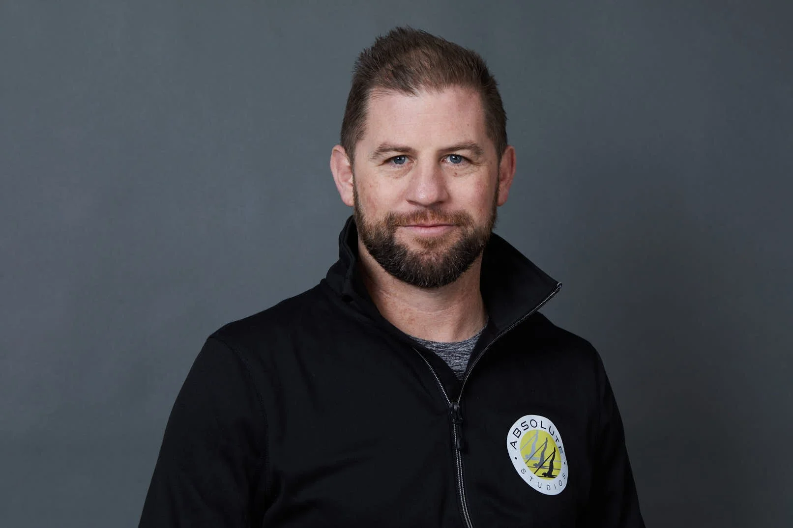A man with a beard and short hair, wearing a black jacket with a logo, standing against a gray background.