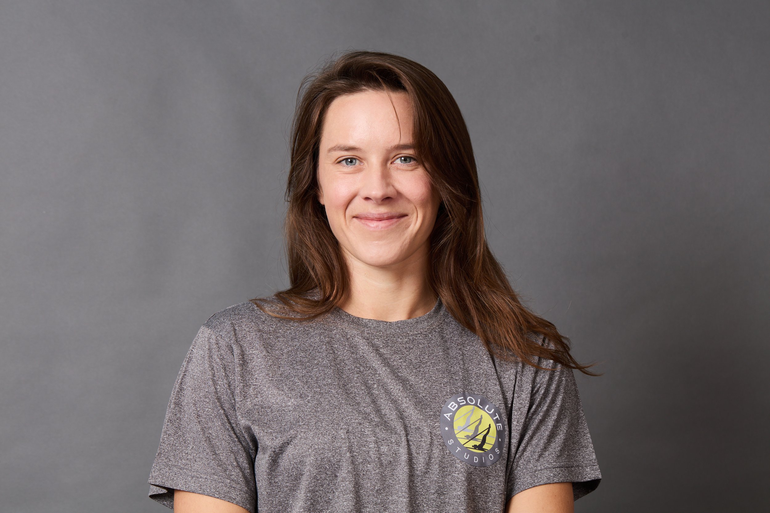 A smiling young woman with shoulder-length brown hair wearing a gray T-shirt with a logo that reads 'Absolute Studios' against a plain gray background.