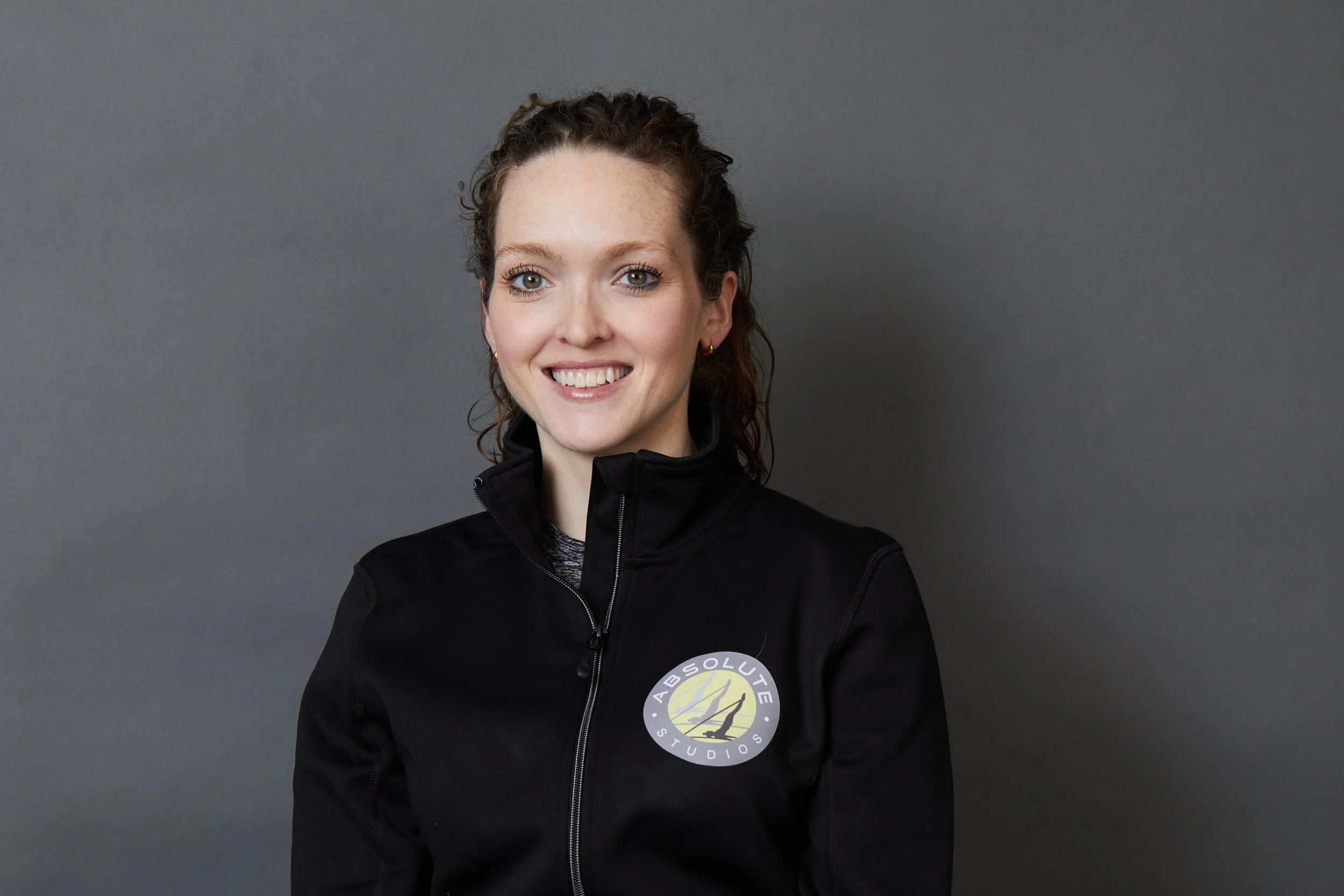A smiling woman with curly brown hair wearing a black jacket with a logo for Absolute Studios on the chest, standing against a plain gray background.