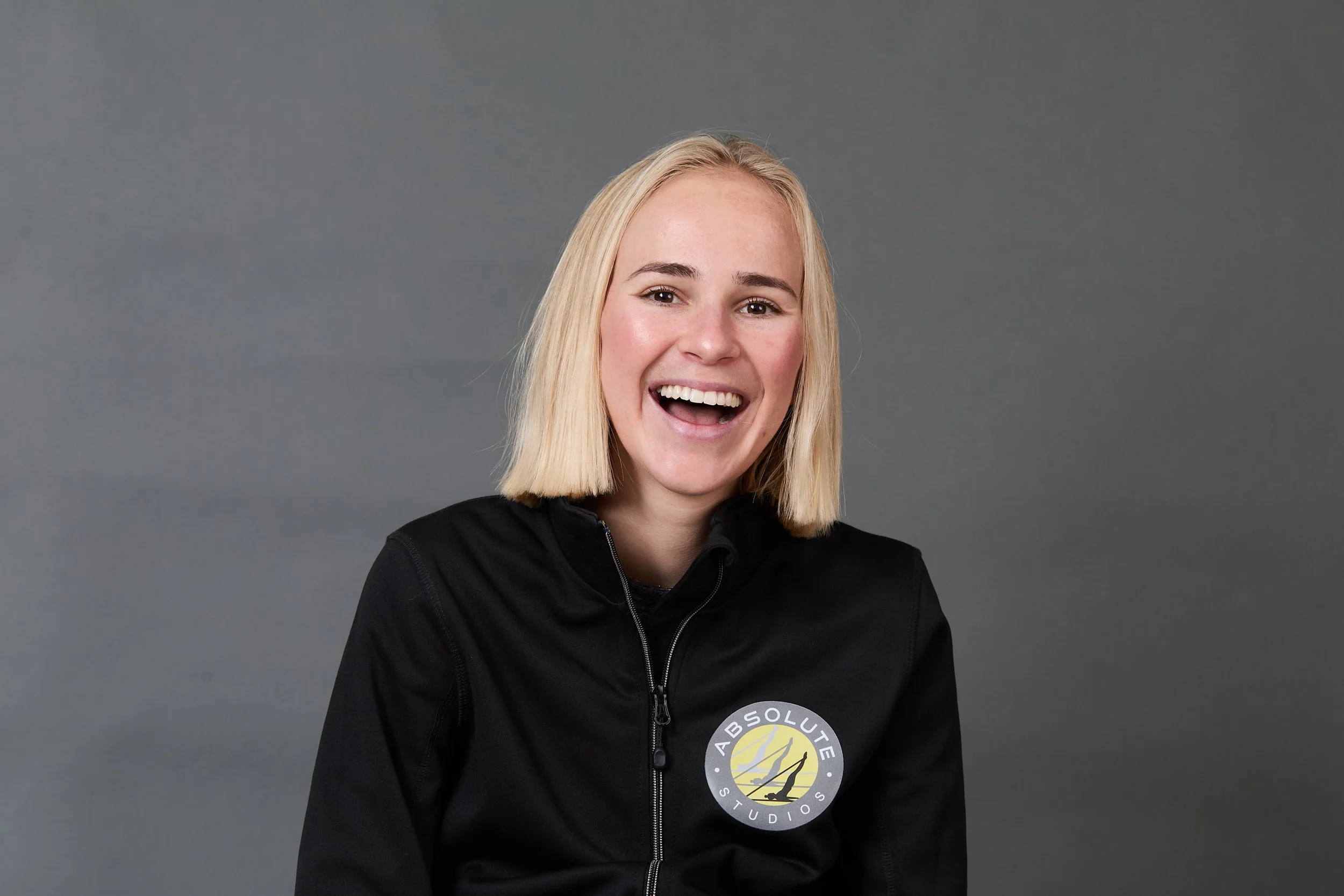 A young woman with shoulder-length blonde hair smiling at the camera, wearing a black jacket with a circular logo that reads "Absolute Studios" on the front, set against a plain gray background.
