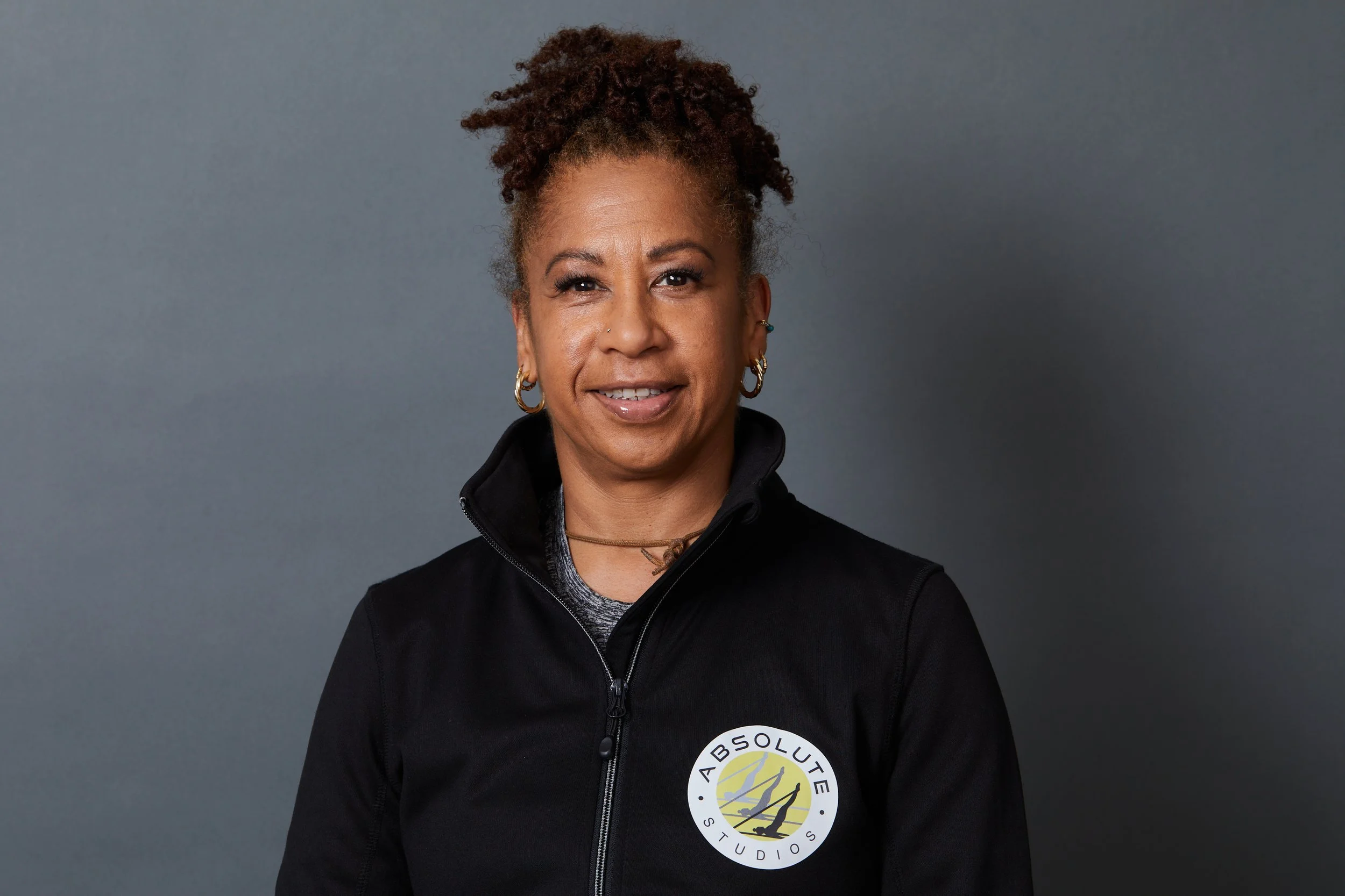 Woman smiling, wearing a black sports jacket with a logo that reads 'Absolute Studios' on the chest, against a gray background.