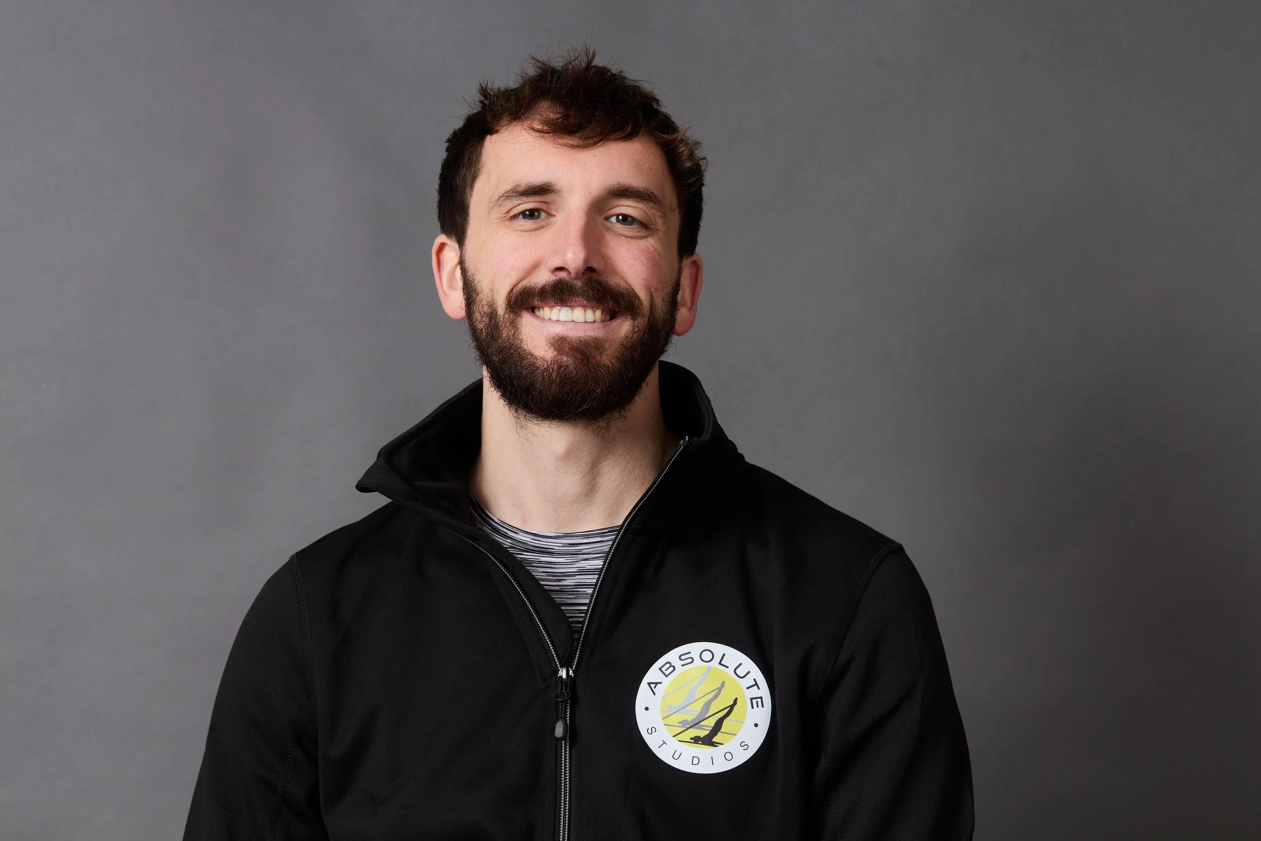 A smiling man with a beard and brown hair, wearing a black jacket with the 'Absolute Studios' logo on it, in front of a gray background.