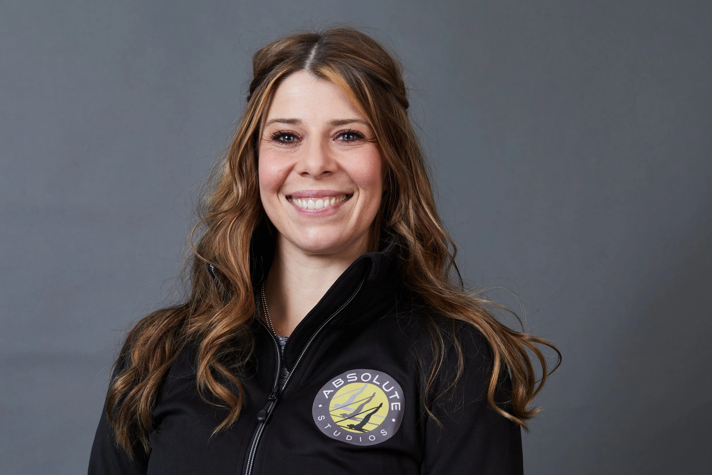 A smiling woman with long wavy brown hair wearing a black zip-up jacket with a logo that reads 'Absolute Studios' on the front, posed against a gray background.