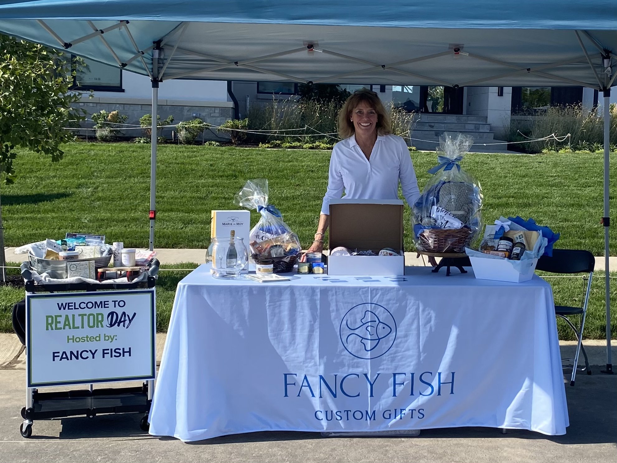 A woman stands behind a table under a canopy at an outdoor event called Realtor Day, hosted by Fancy Fish, displaying various gift items and promotional materials.