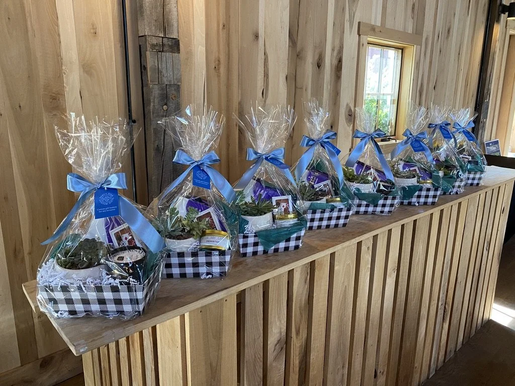 A row of gift baskets wrapped in clear cellophane with blue ribbons, sitting on a wooden counter inside a rustic wooden room.