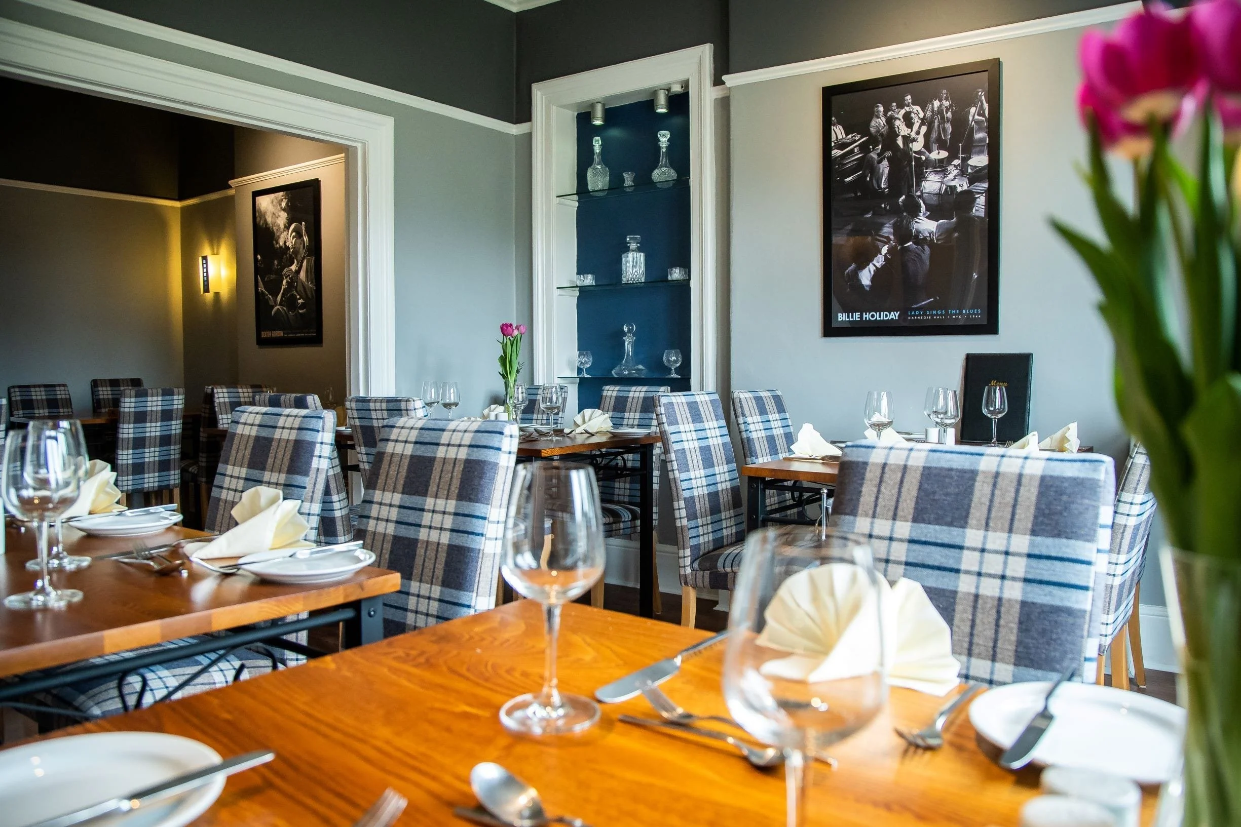 Elegant dining room with wooden tables, checkered upholstered chairs, wine glasses, folded napkins, and a vase with pink tulips. The wall features a large black and white poster of Billie Holiday and artwork of jazz musicians.