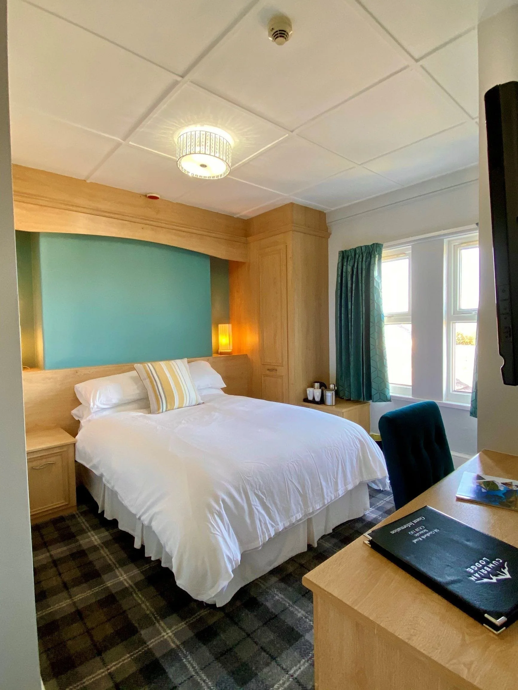 A neatly made hotel room with a white bed, a blue accent wall behind the bed, wooden furniture including a wardrobe and nightstands, and windows with teal curtains letting in natural light.
