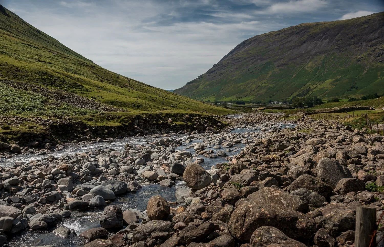 A mountain valley with a rocky stream flowing through it, green slopes on either side, and a partly cloudy sky overhead.