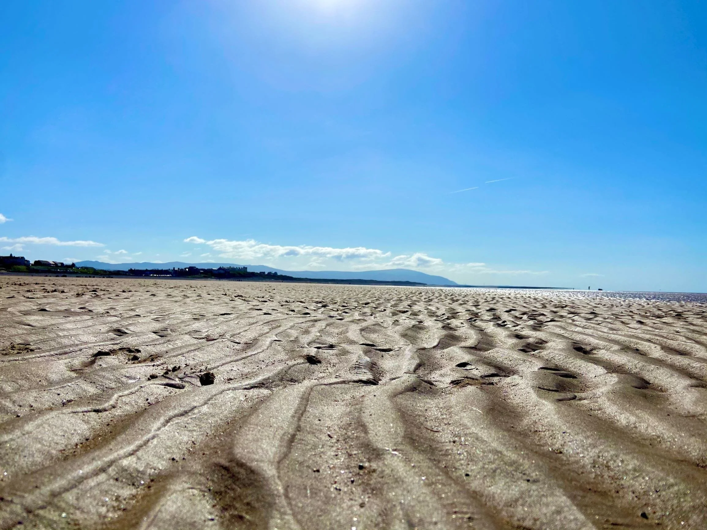 Empty sandy beach with rippled patterns, distant houses, and a bright blue sky with a few clouds