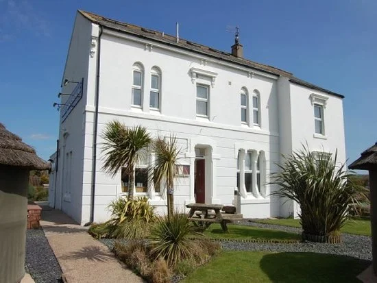 A white two-story house with multiple windows, surrounded by a garden with grass, palm trees, and a picnic table, under a clear blue sky.