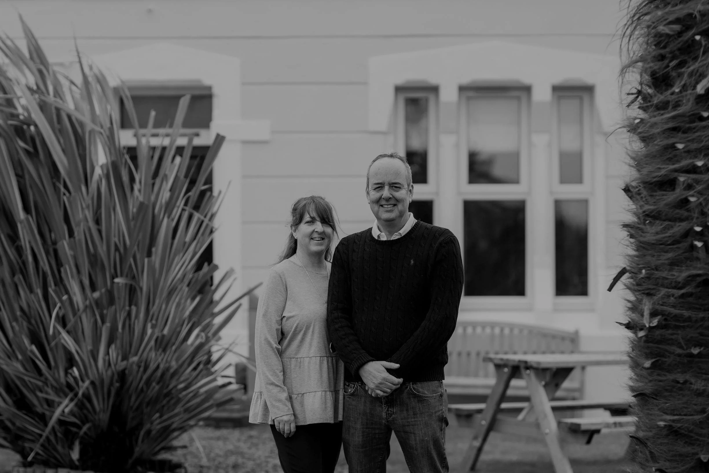 A smiling man and woman standing outside in front of a house with large windows, surrounded by bushes and a picnic table.