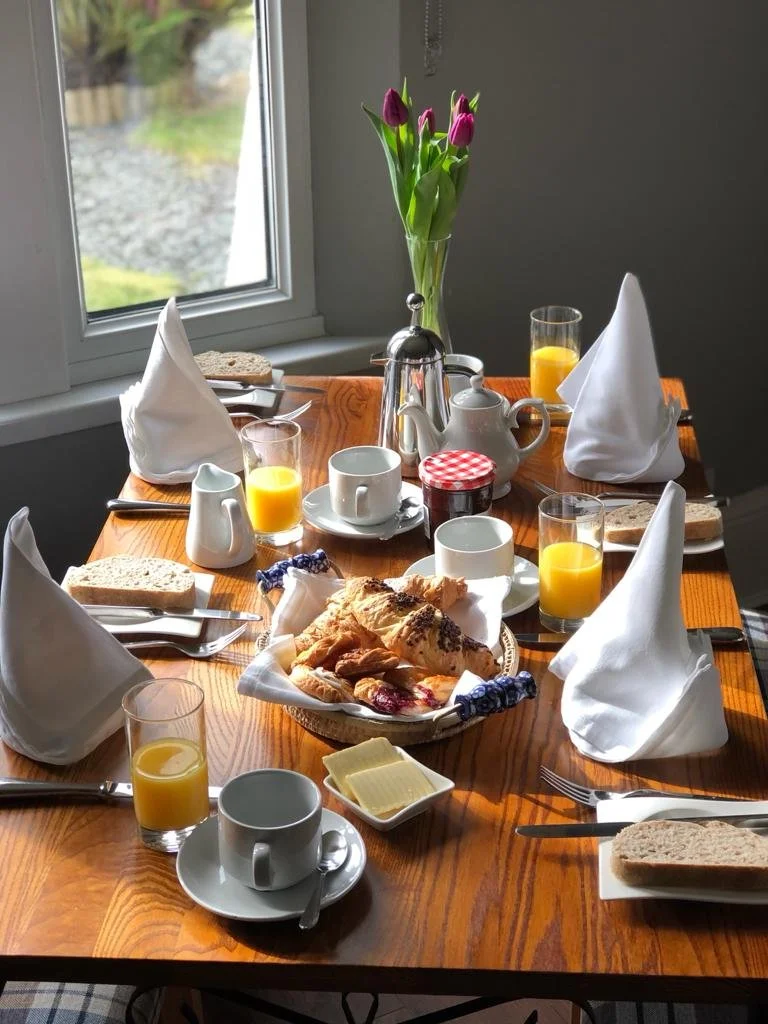 A breakfast table set near a window with six plates, cups, and glasses, including croissants, bread, butter, jam, orange juice, and a vase with purple tulips.