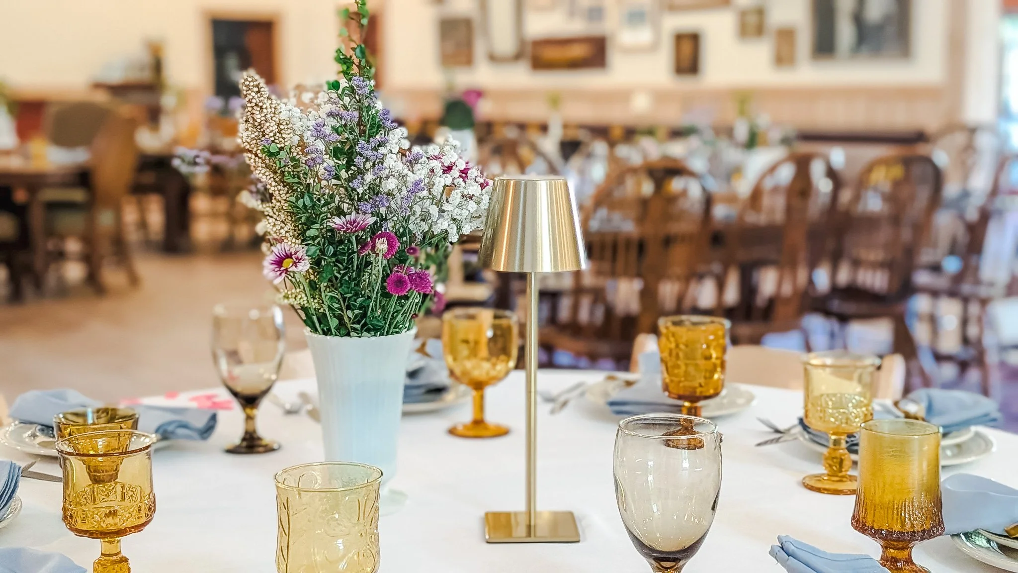 A table set for a meal with a large white vase of mixed flowers, a small gold lamp, and amber-colored glasses, in a decorated dining area.