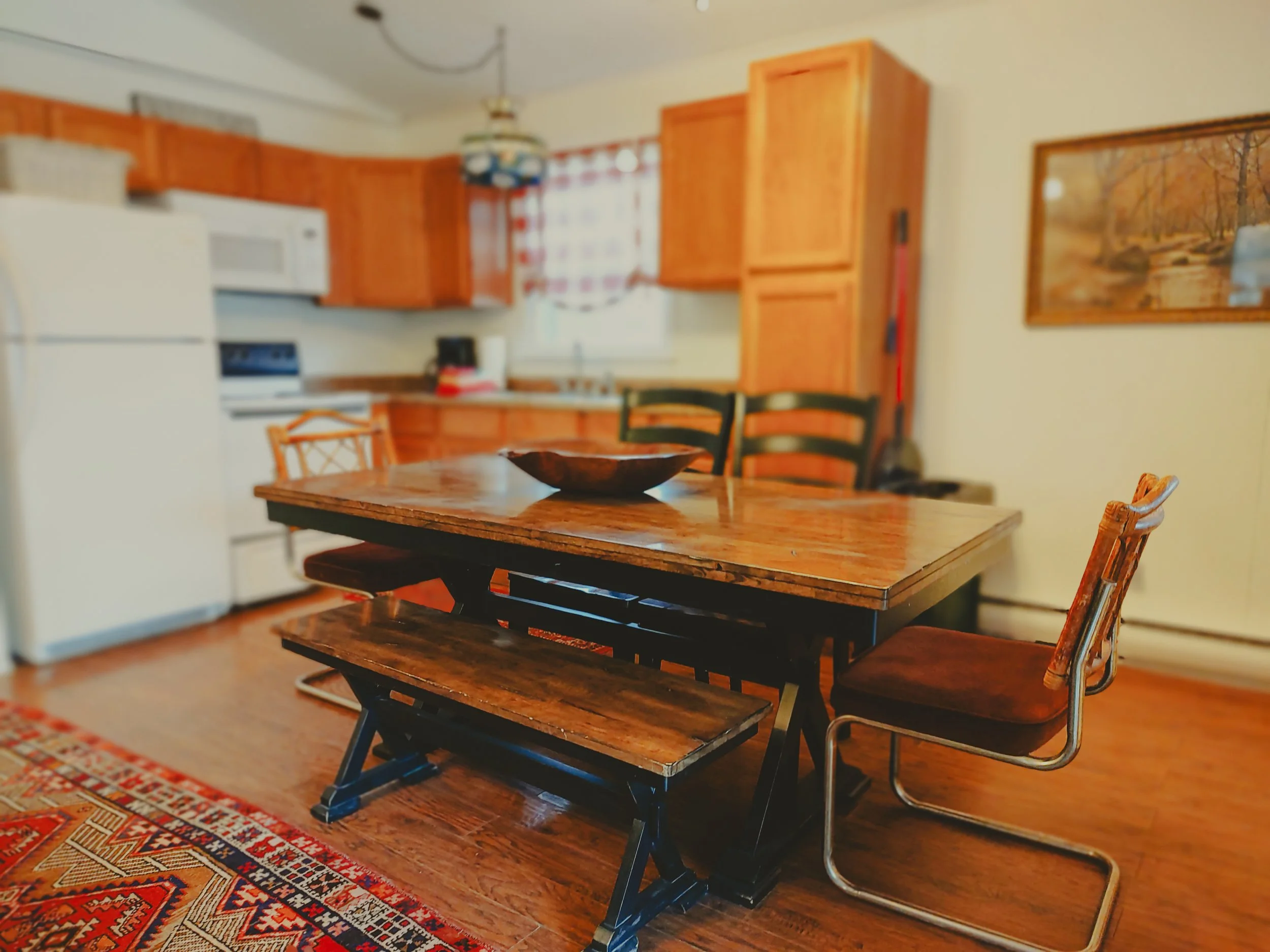 A dining area with a wooden table, a bench, and chairs in a kitchen with wooden cabinets, white refrigerator, stove, and microwave, with a colorful rug on a wooden floor and a framed landscape picture on the wall.