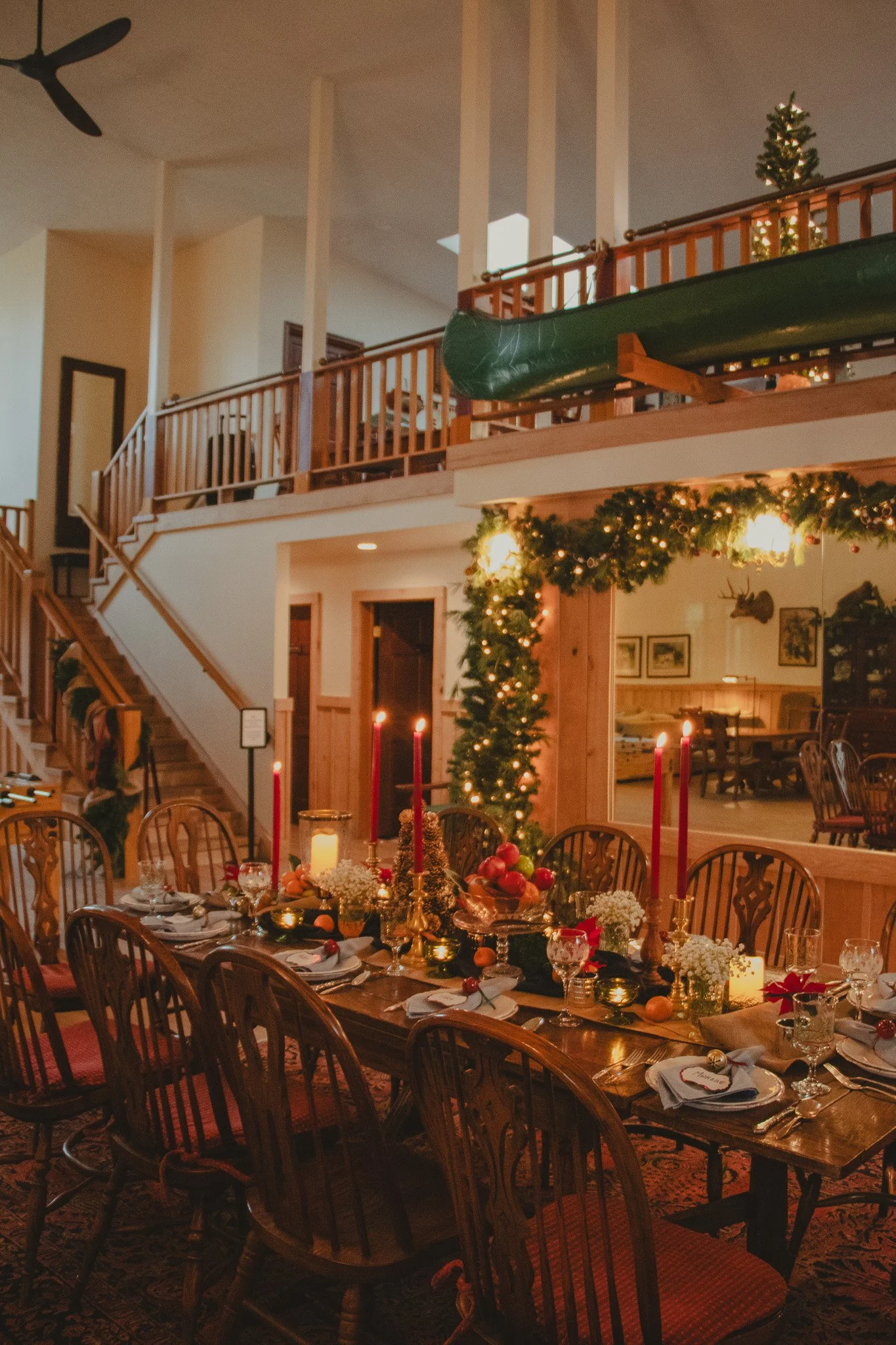 A festively decorated dining area with a wooden table set for Christmas, featuring red candles, Christmas ornaments, and flowers, illuminated by warm lights, with a garland overhead, and a large window with a view into a cozy room