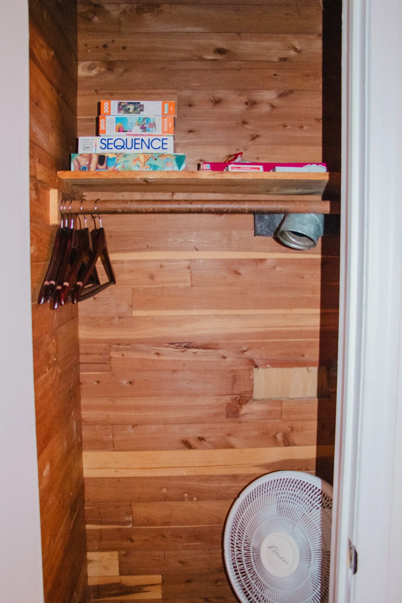 Small wooden closet with a shelf holding a puzzle, a game box, and other small items. Hangers are on the left side, and a white electric fan is on the floor in front of the closet.