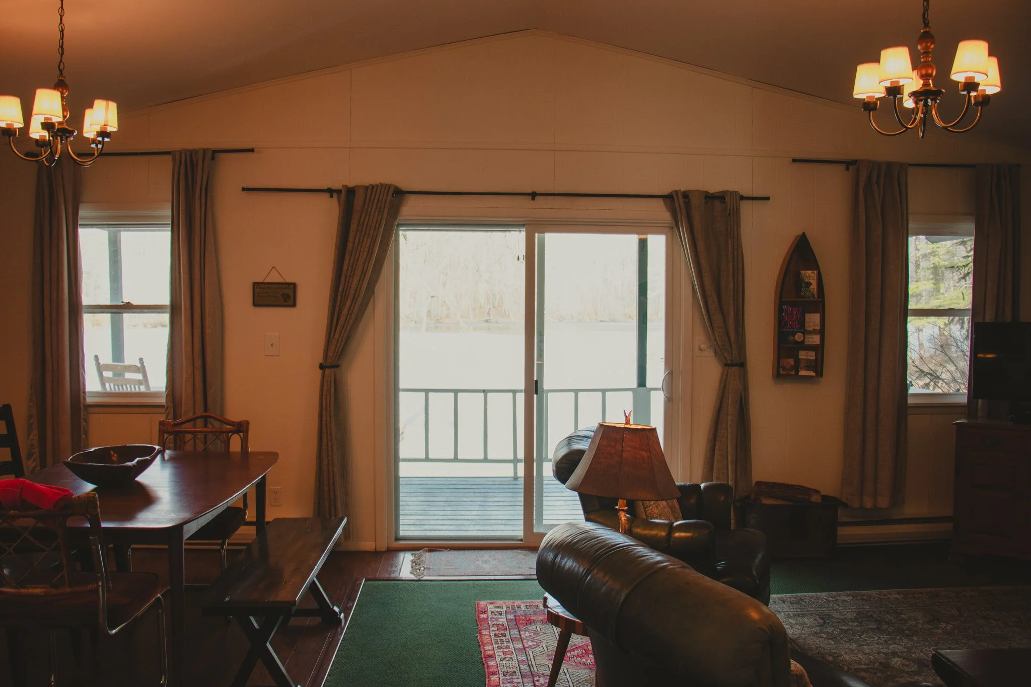 Living room with wooden table, leather chairs, sliding glass door leading to balcony, windows with curtains, ceiling lights, and a television.