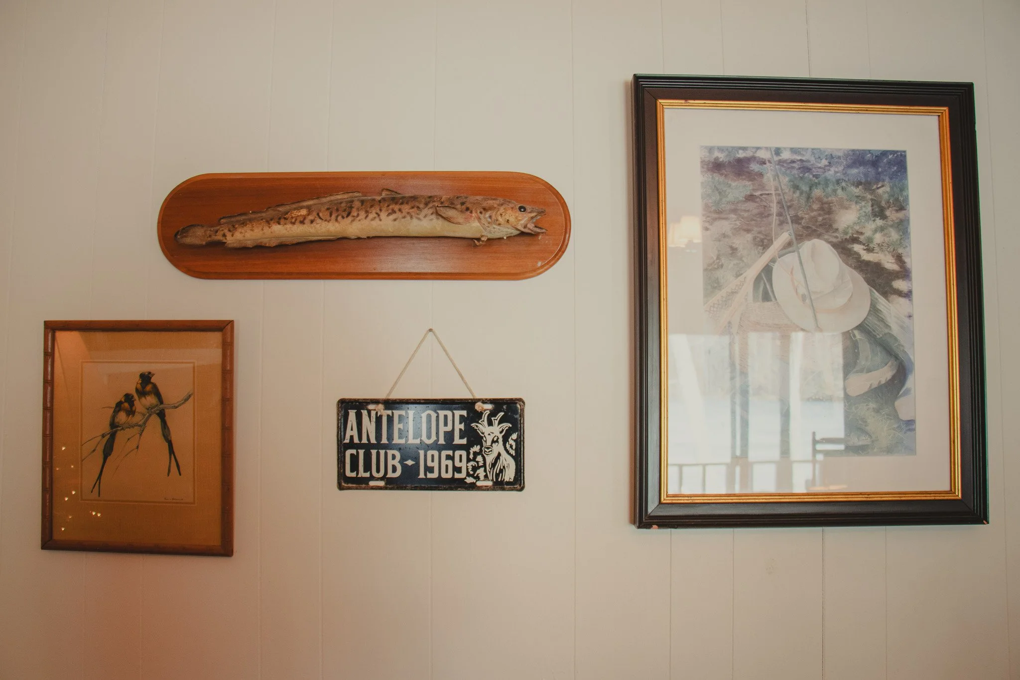 Wall decorated with a mounted fish on a wooden plaque, a framed picture of two birds, a sign reading 'ANTELPE CLUB - 1969', and a framed painting of a white hat and fishing rod.