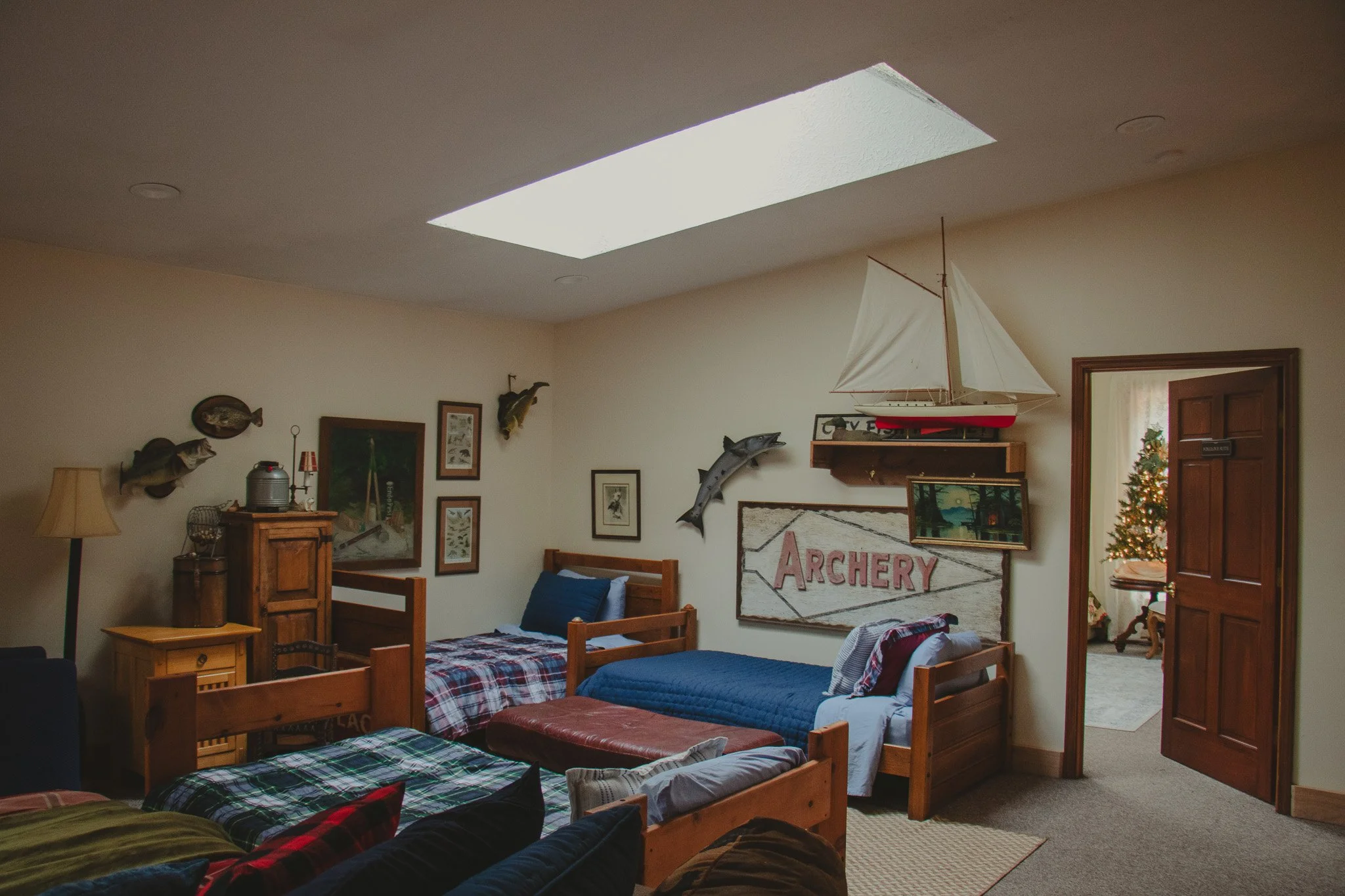 A bedroom decorated with fishing and archery themes, featuring three beds with plaid and solid bedding, a Christmas tree visible through a doorway, and a large skylight in the ceiling.