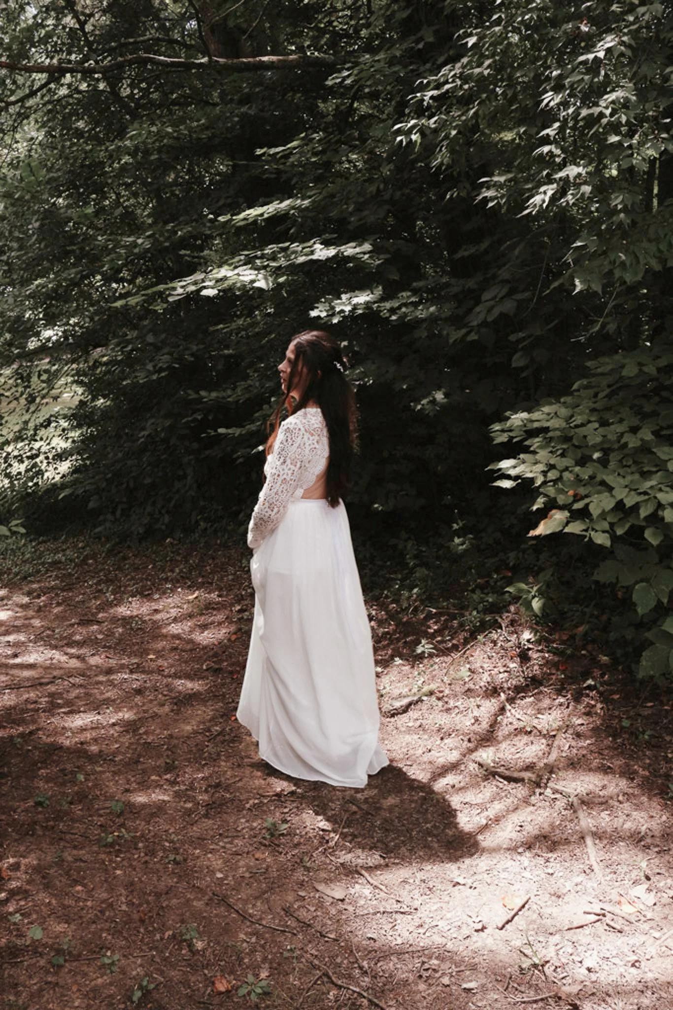 A woman wearing a white dress with lace top standing on a dirt path in a wooded area with dense greenery.
