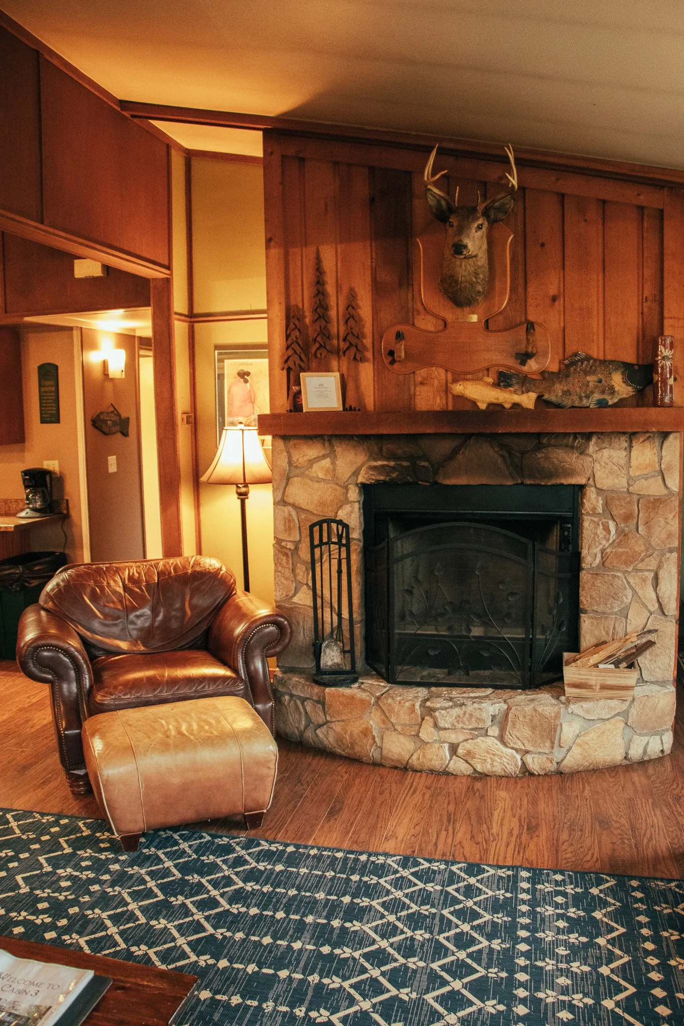 Cozy living room with a stone fireplace, mounted deer head, and wood-paneled wall. A leather armchair with an ottoman is near the fireplace, with a patterned rug on the wooden floor.