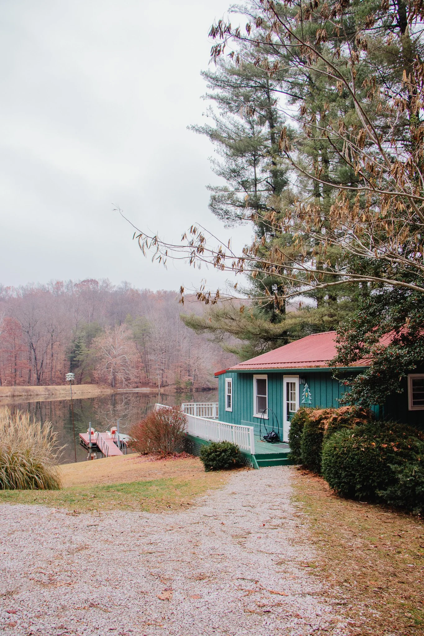 A teal house with a red roof beside a lake, surrounded by trees with autumn and winter foliage, some bushes, and a gravel driveway leading to the house.