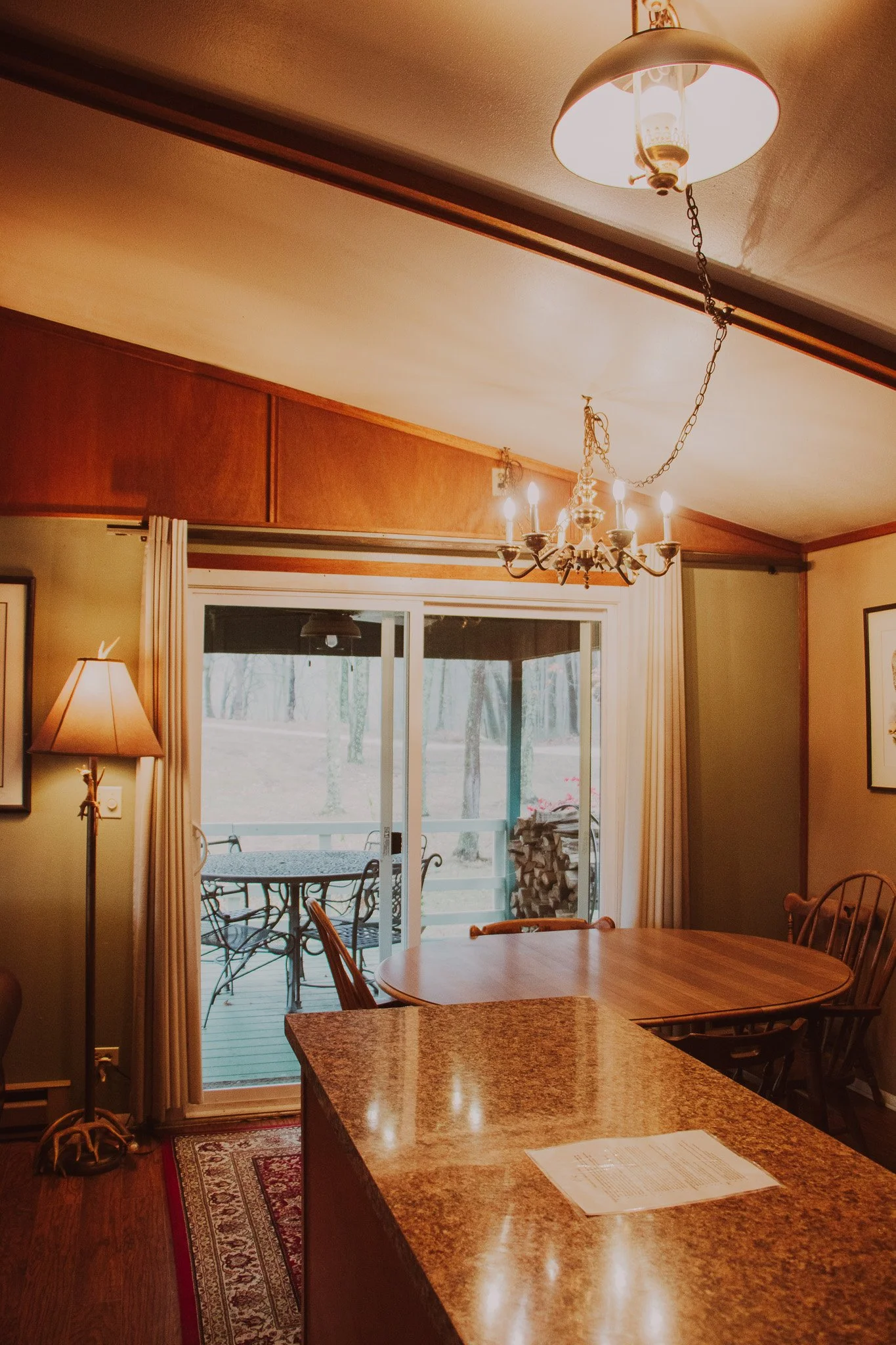 Interior view of a dining room featuring a wooden table, a granite kitchen counter, a sliding glass door leading to a deck outside, and a chandelier hanging from the ceiling.