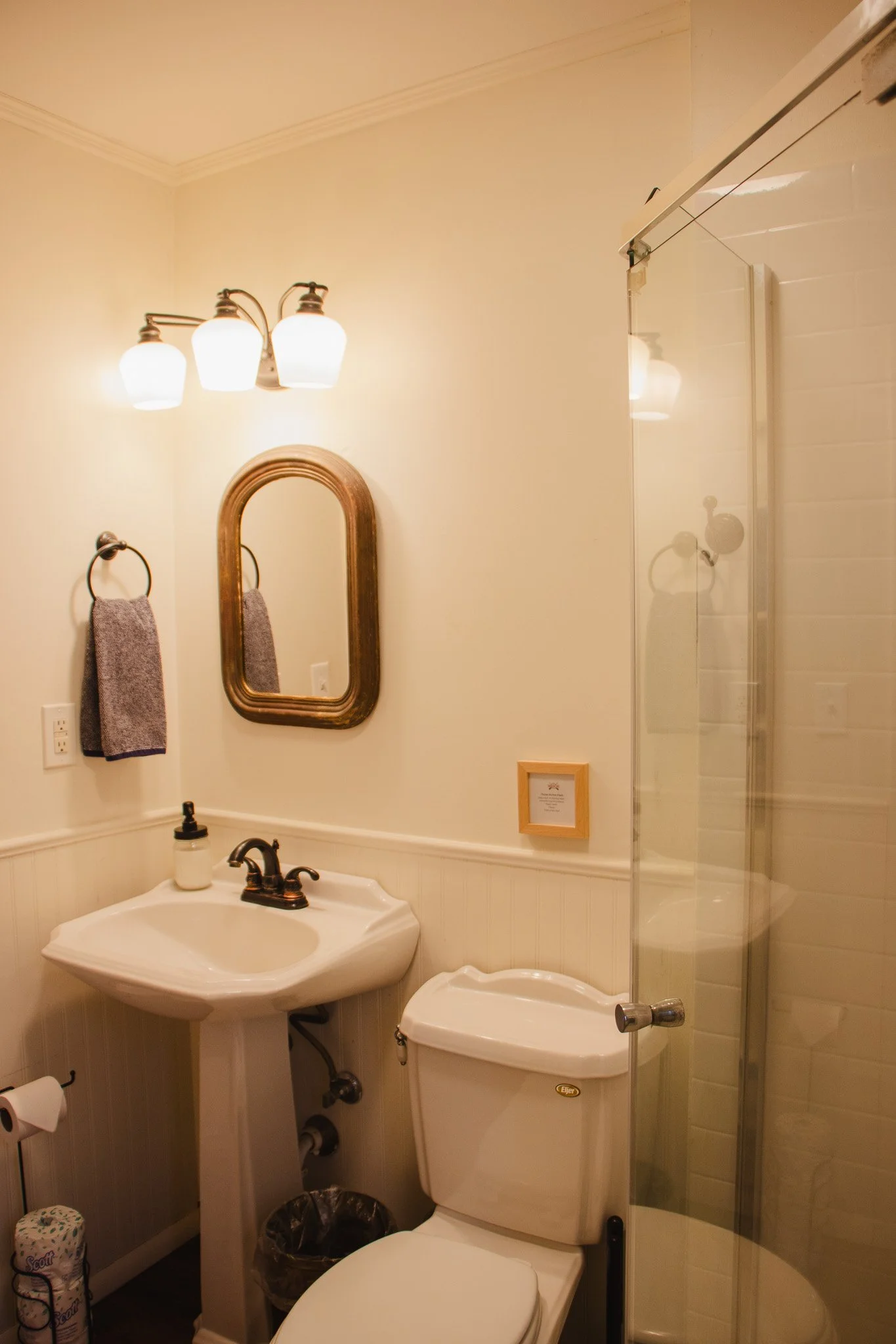 A small bathroom with a white sink and a oval mirror, a toilet, a framed picture on the wall, a towel ring with a towel, and a glass shower enclosure with a door.