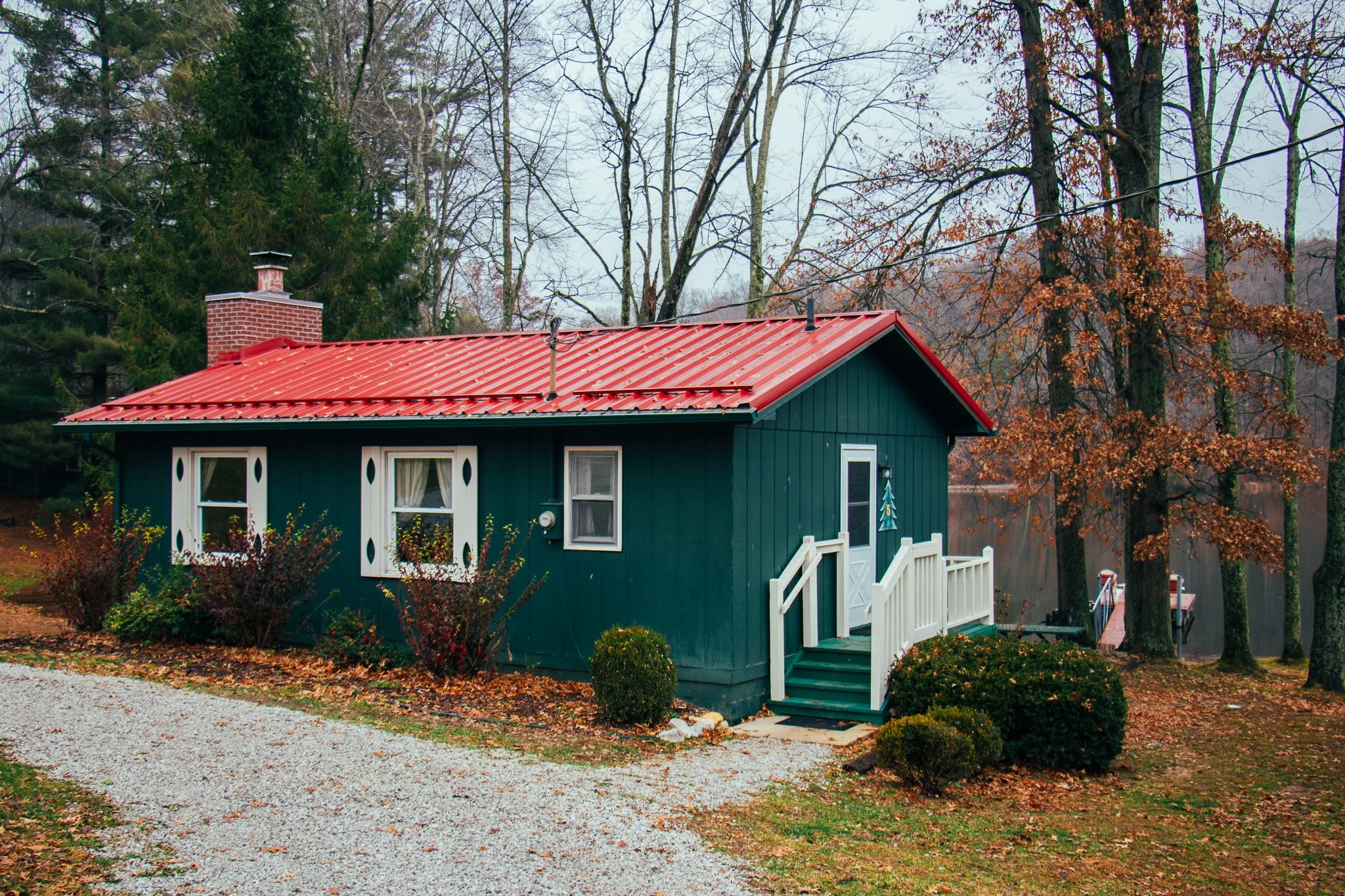 A small teal house with a red metal roof, white trimmed windows, and a white staircase with a door surrounded by trees with autumn leaves.