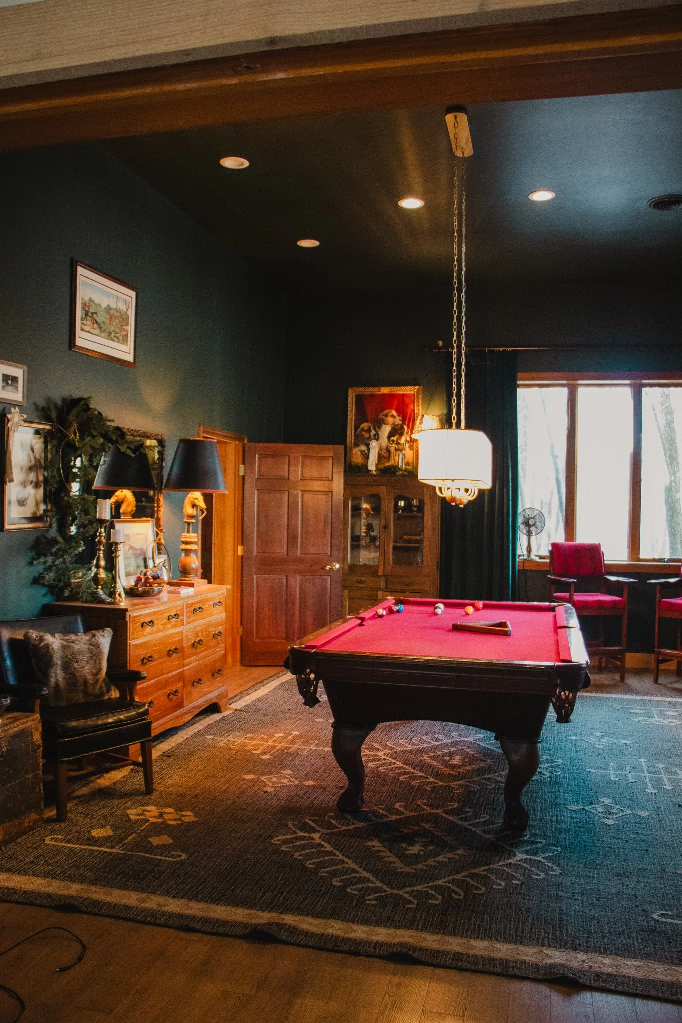Interior of a living room with a pool table, dark green walls, wooden furniture, artwork, and a window with curtains.