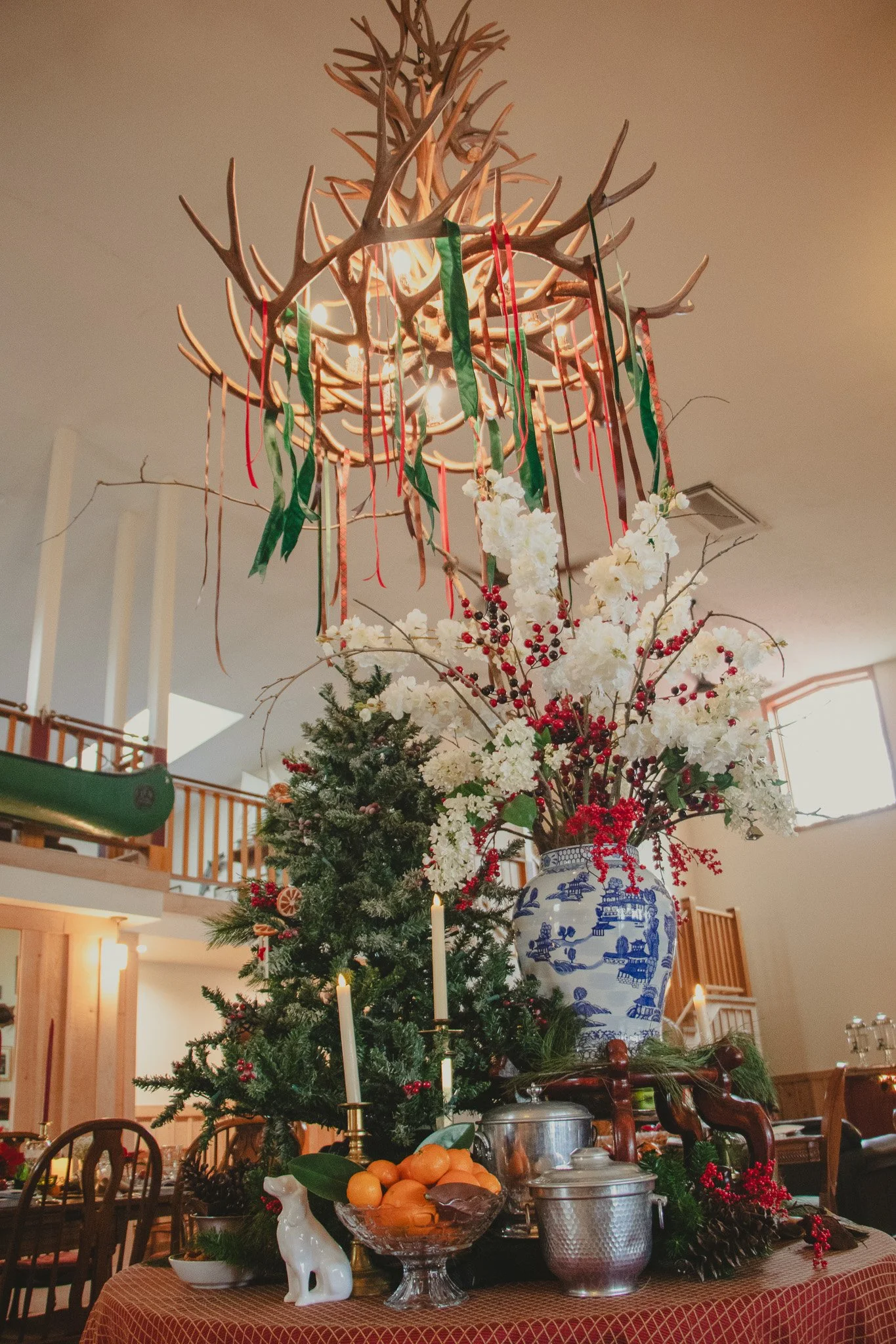 A festive holiday table decorated with a large blue and white porcelain vase filled with white and red flowers, a Christmas tree, candles, and food. A chandelier made of antlers hangs above.