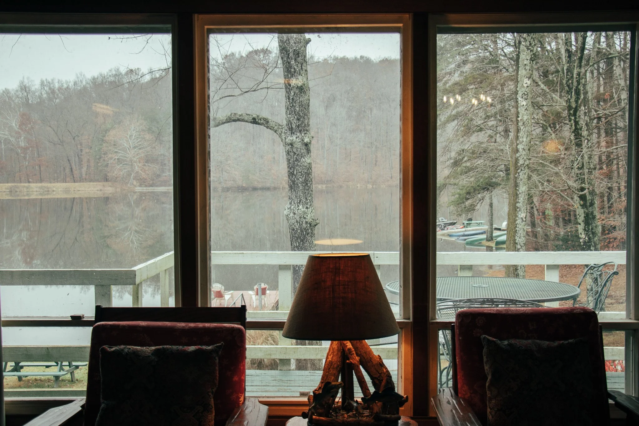 Interior of a cozy room looking out a window at a wooded lake scene with trees and boats, a table, chairs, and a lamp inside.