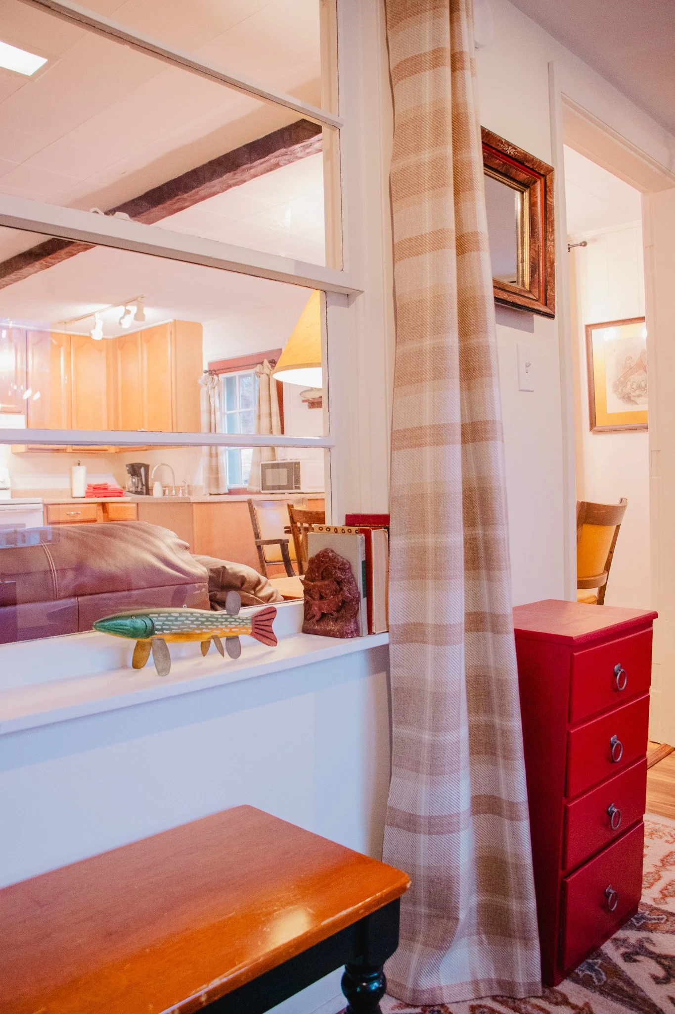 View into a living room and kitchen area through a large window with a checkered curtain, displaying a red chest of drawers, a table, and various decor items.