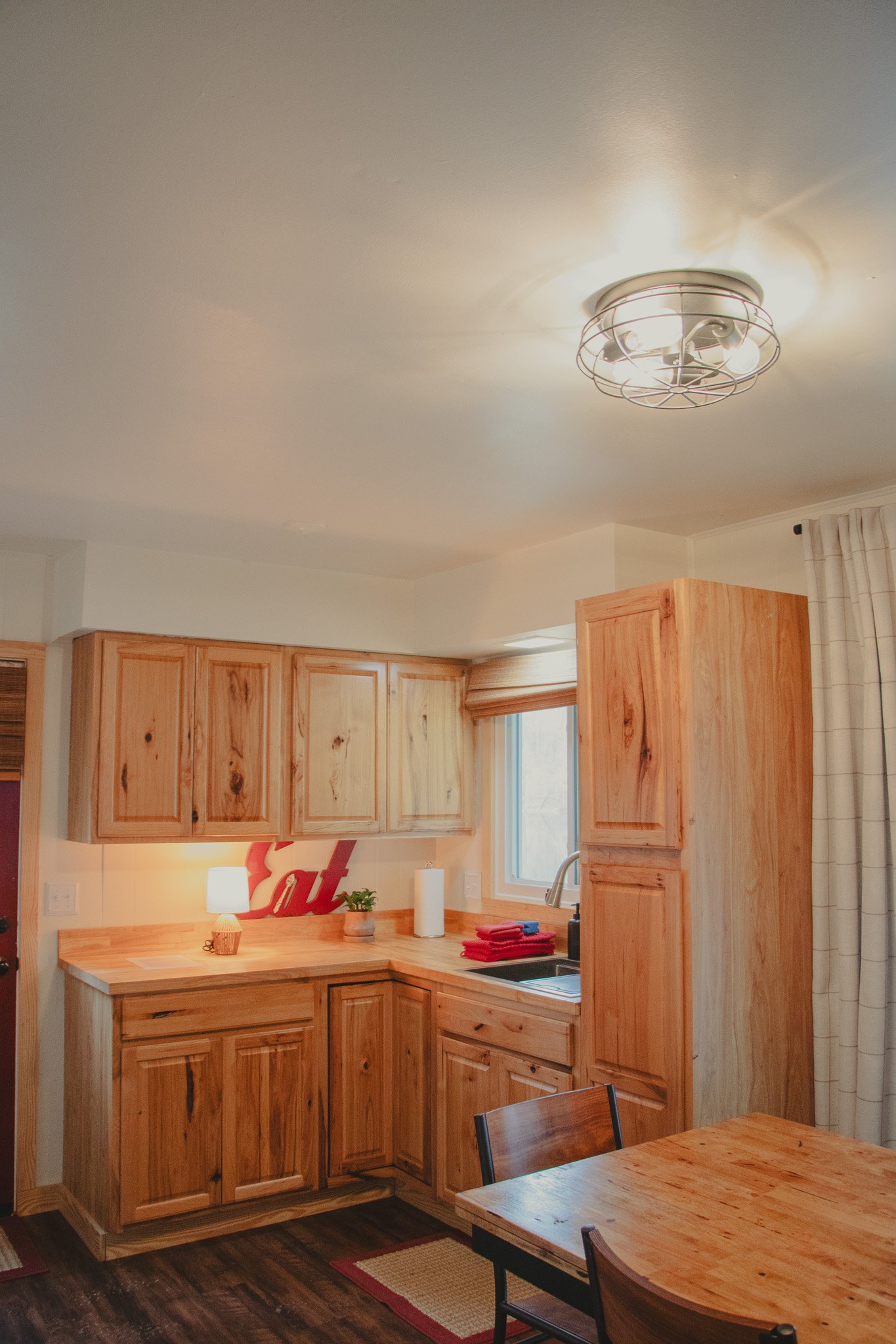 Kitchen with wooden cabinets, a window, a sink, a small lamp, a paper towel roll, and red cloths on the counter. Part of a dining table with chairs is visible.