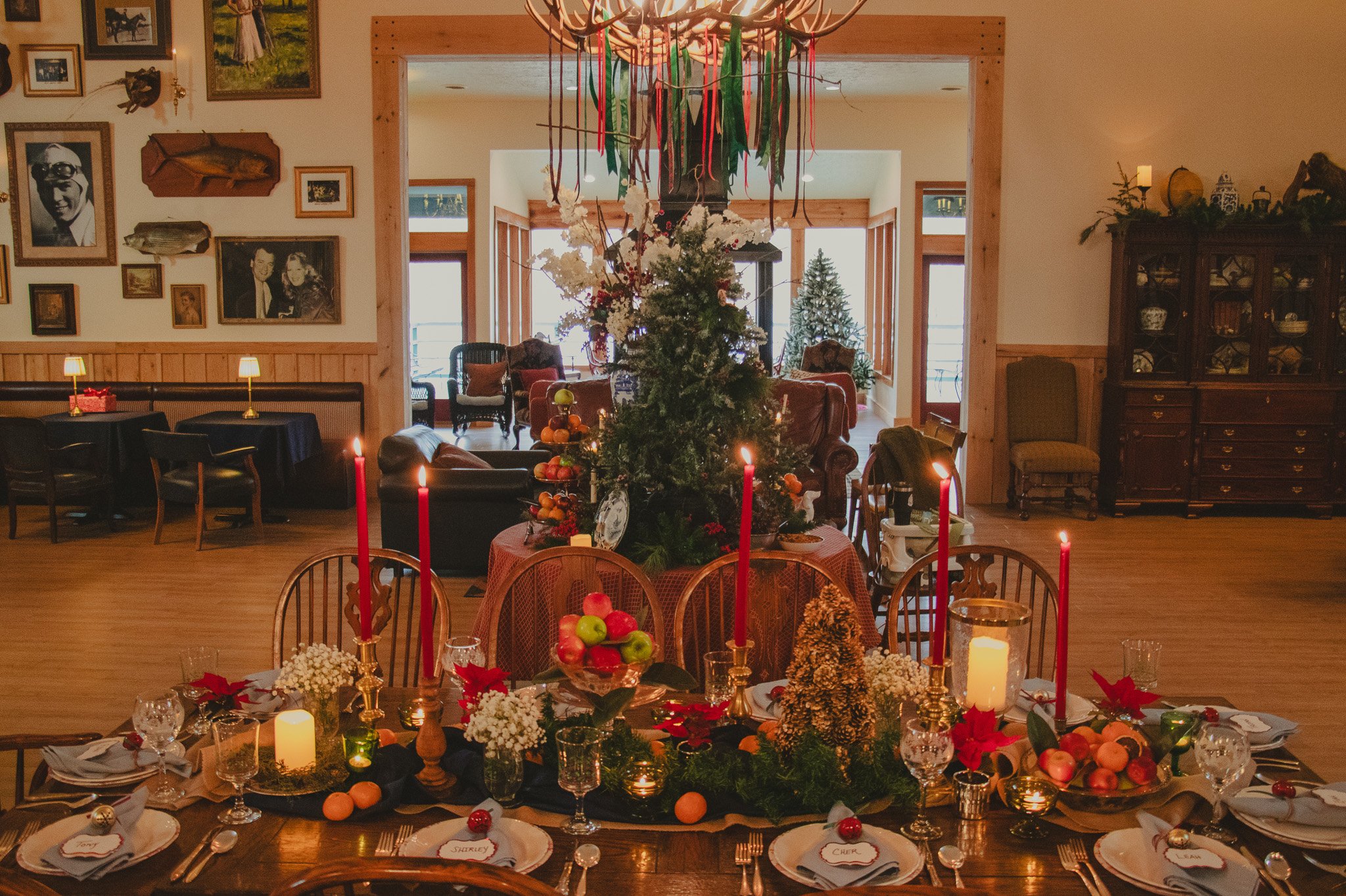 A decorated dining table for Christmas with candles, flowers, and apples in a cozy, festively decorated room with a Christmas tree in the background.