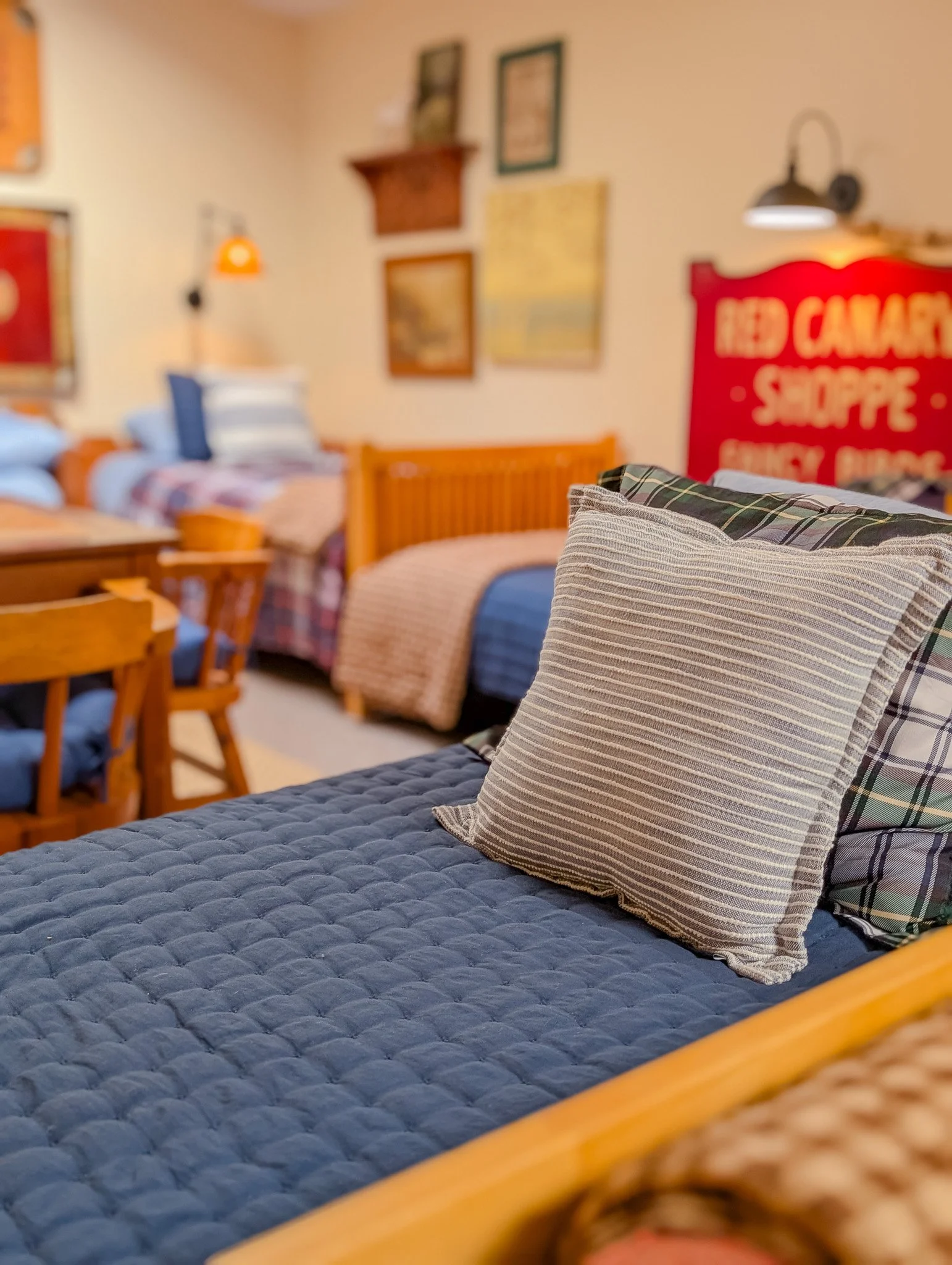 Close-up of a plaid pillow on a blue quilted bedspread in a cozy bedroom with vintage decor, including framed pictures on the wall and a red sign in the background.