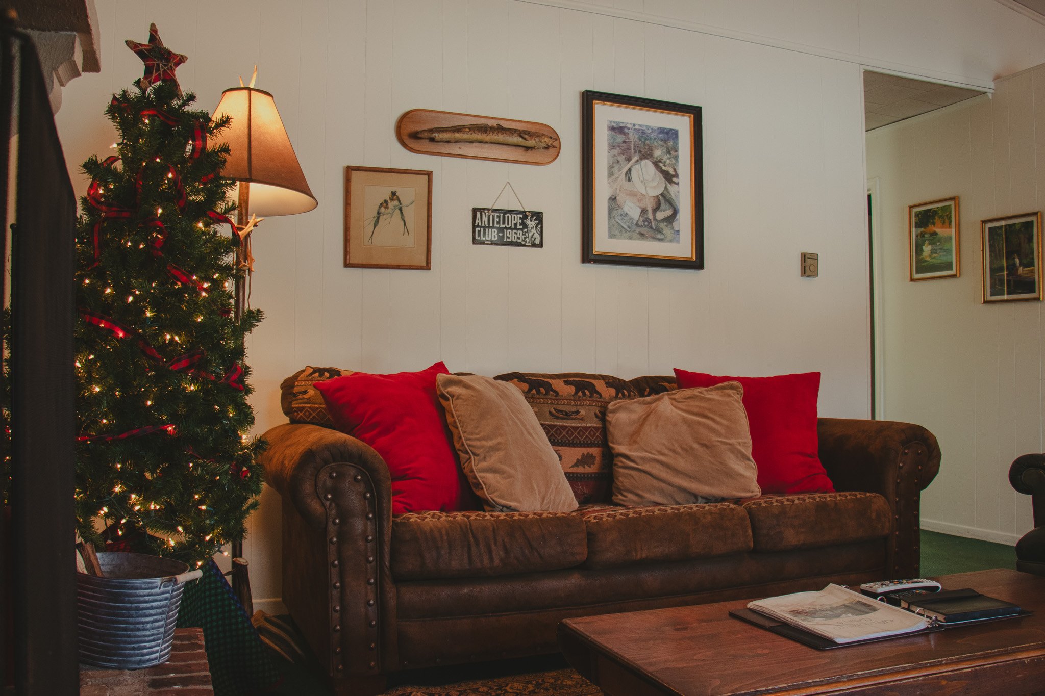 Living room with decorated Christmas tree, front of a brown sofa with red and beige pillows, wall decor including framed pictures and wooden plaques, and a wooden coffee table.