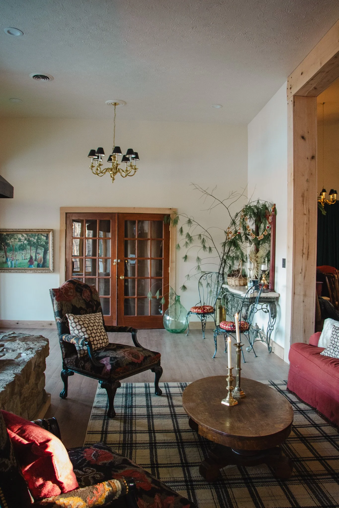 A cozy living room with vintage furniture, including a floral armchair, a red sofa, and a wooden coffee table with brass candlesticks. There is a chandelier hanging from the ceiling, a painting on the wall, and decorative plants near a glass-paneled 
